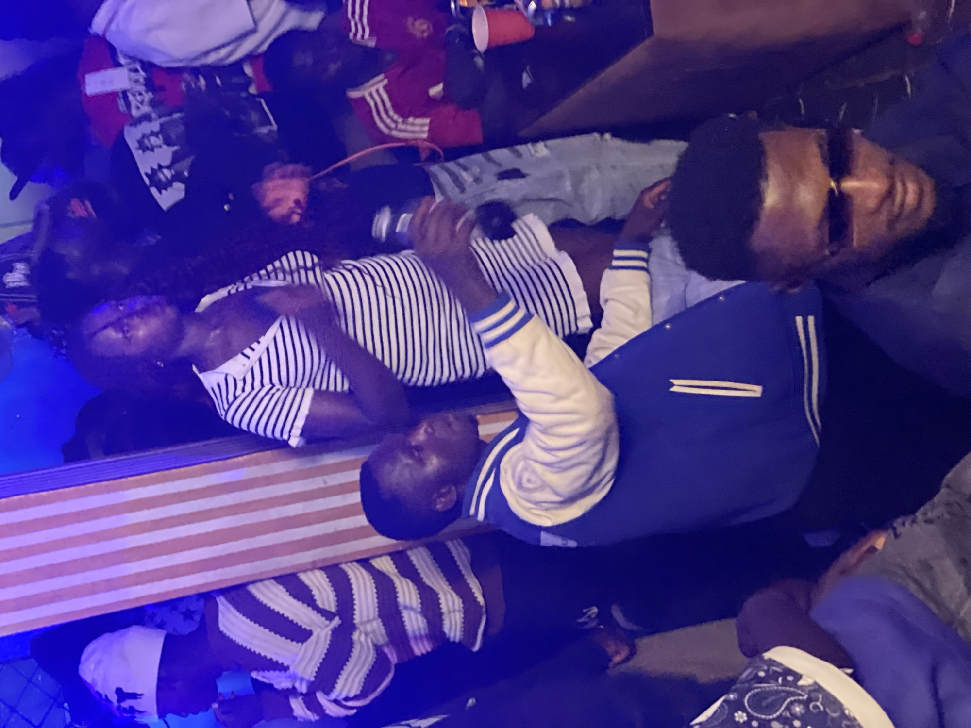 In Wumat, Nigeria, a group of people are gathered indoors, possibly at a bar or club. The lighting is dim, with blue and pink hues creating a vibrant atmosphere. In the foreground, a man wearing sunglasses and a dark shirt is looking down, while a man in a blue and cream jacket is holding a bottle, seemingly interacting with someone to his right. To the left, a person in a striped sweater and beanie is visible, and a woman in a striped top is standing further back. The environment appears crowded, with hints of other patrons and objects like red cups on a counter.