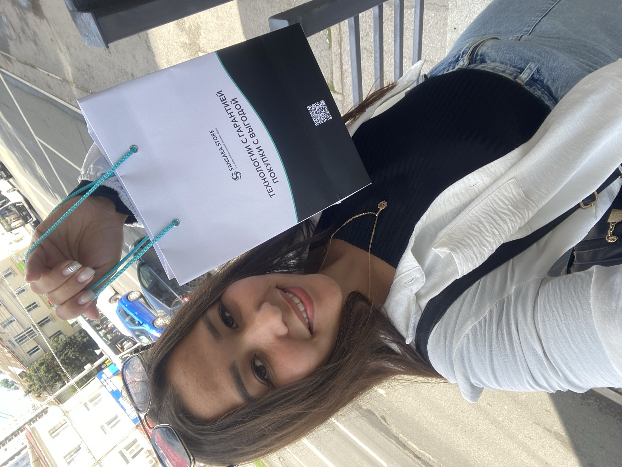 A young woman with long, brown hair, wearing a black top, a white unbuttoned shirt, and blue jeans, is taking a selfie outdoors in the city of Ufa, Russia. She is holding up a white shopping bag with teal handles, which has the logo "SANSARA STORE" and text in Russian that translates to "TECHNOLOGIES WITH GUARANTEE, PURCHASES WITH BENEFIT." A QR code is visible on the bag. Sunglasses are perched on her head, and she is wearing a gold necklace. Behind her, a street scene with buildings, cars, and a bus is visible. The lighting suggests it is daytime.