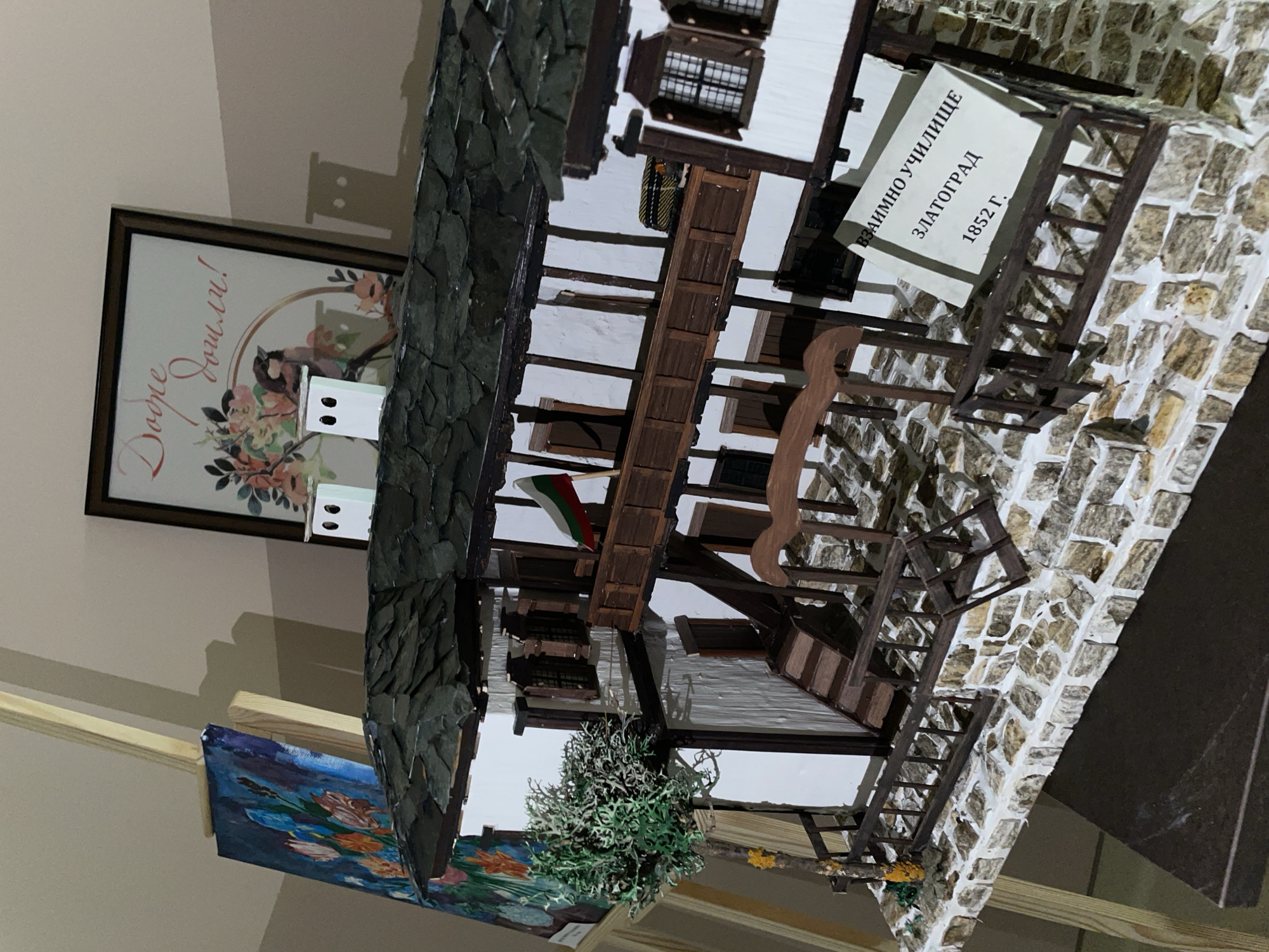 The image shows a diorama or model of the Vzaimno Uchilishte in Zlatograd, Bulgaria, established in 1852. The model features a stone facade, white walls, and wooden details. A sign indicates the name and founding year of the school. The model is situated near other objects: a framed picture and a blue-toned painting. The background is a beige wall.