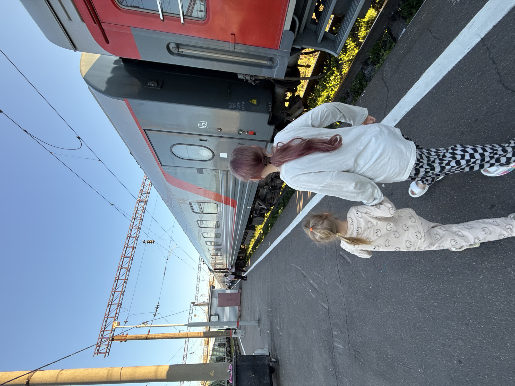 In Uzunovo, Russia, two young girls walk along a train platform beside a train. The girl in the foreground wears a light-colored pajama set with a bear print, while the girl in front of her has pink-tinged hair and wears a white long-sleeved shirt with black and white checkered pants. They are walking away from the camera, holding hands. The train, a passenger car with gray and red accents, is parked on the tracks. Overhead, power lines and supporting structures are visible against a clear blue sky. The platform is paved with asphalt, marked by a white line along the edge. A small utilitarian building and a trash bin are also present on the platform. The scene appears to be a train station.