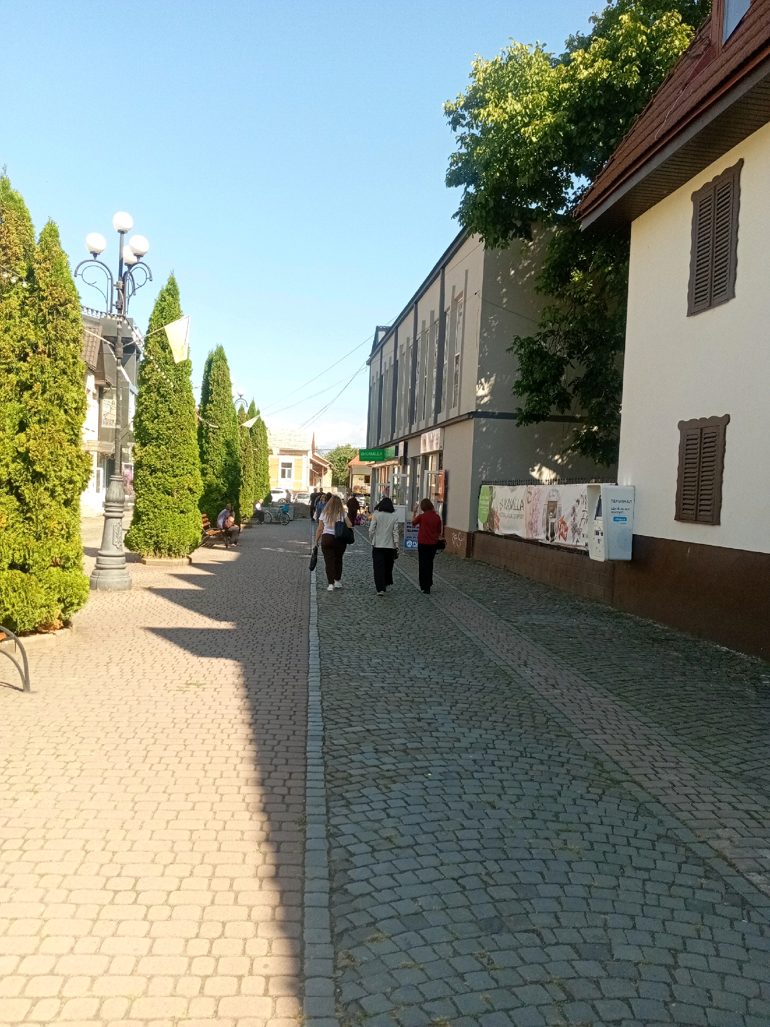 The image shows a paved street in Tiachiv, Ukraine, on a sunny day. The street is lined with tall, conical green trees on the left and buildings on the right. The paving stones are of two colors: light beige on the left and dark gray on the right. Three women are walking away from the camera on the right side of the street. One woman in the middle is wearing a white shirt and brown pants. The woman to her left is wearing a white shirt and dark pants, and the woman to her right is wearing a dark shirt and pants. A man is sitting on a bench on the left side of the street, and another man is visible in the background near a bicycle. A lamppost with three lights stands on the left side of the street, near the trees. There are signs and advertisements on the buildings. The sky is clear and blue.