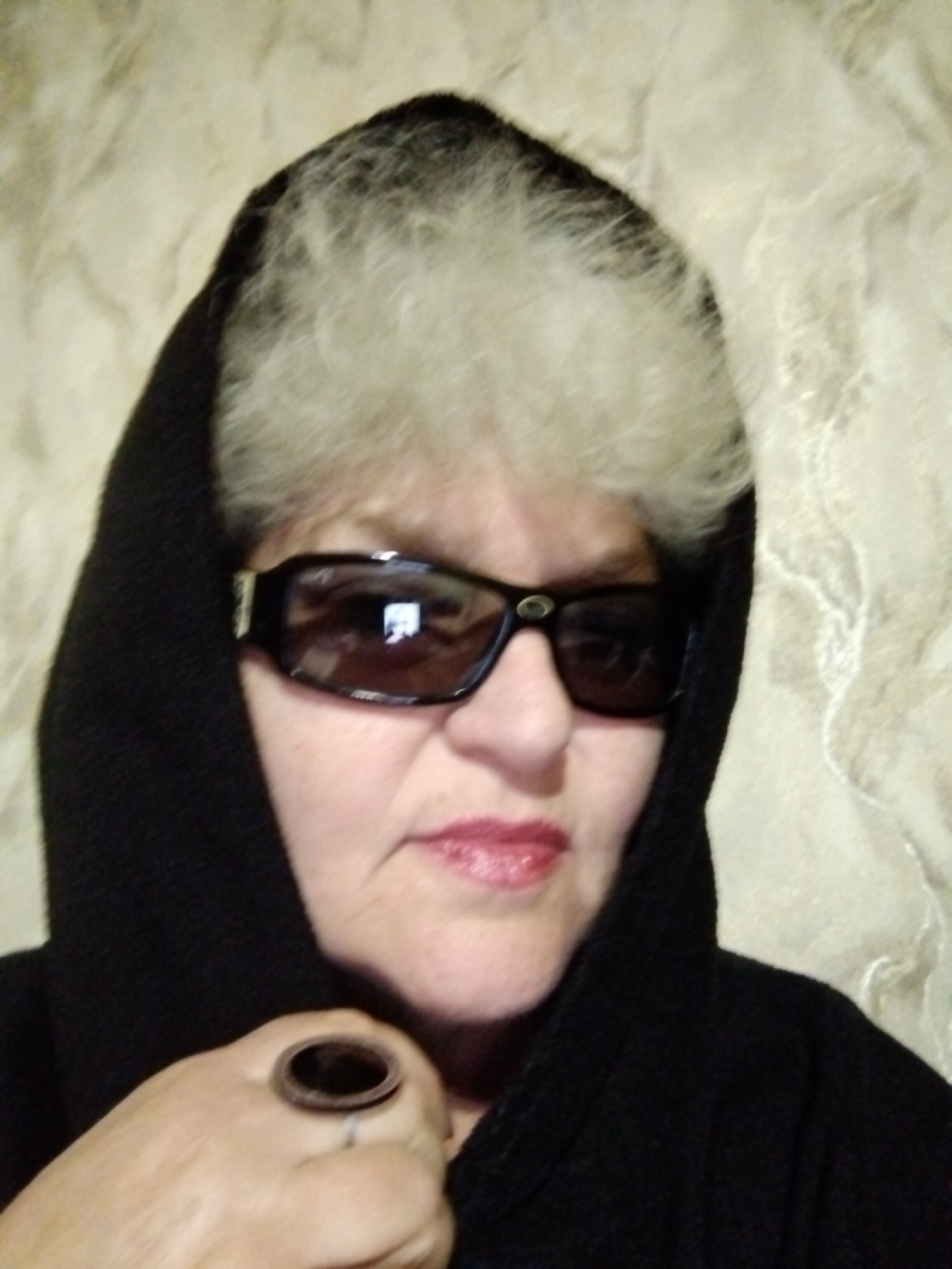 A mature woman is photographed from the chest up, wearing a black hooded garment and large, dark-lensed sunglasses. Her hair is short, curly, and silver-gray. She has fair skin and wears red lipstick. A large ring with a dark, oval stone adorns her left hand. The background is a textured, off-white or beige surface. There is no visible indication of the city of Horishni Plavni, Ukraine.