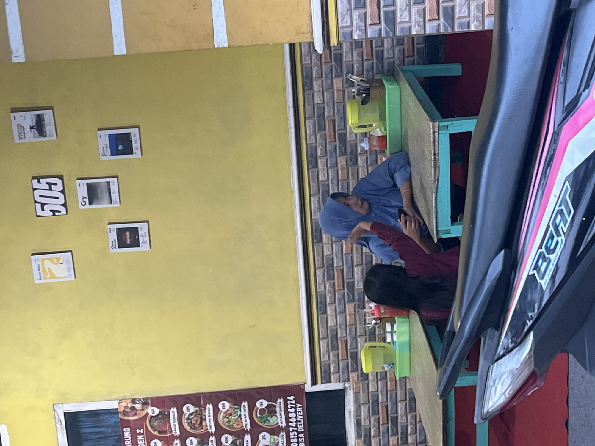 The image captures a scene at a restaurant in Margodadi, Indonesia. Two individuals, a woman wearing a blue hijab and a younger person with long dark hair, are seated at a table. The woman is looking downwards, while the younger person appears to be gesturing. On the table are two green trays, one containing a yellow jug and cutlery, and the other holding a bottle of sauce. The restaurant's yellow wall is decorated with framed posters and the number "505." A menu board with various food items and prices is visible to the left. The overall environment suggests a casual dining setting.