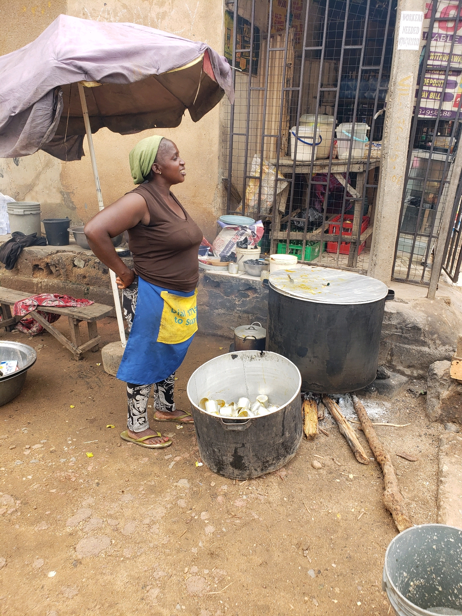 A Black woman wearing a green headscarf, a brown sleeveless shirt, a blue apron with a yellow pocket, and patterned leggings stands outdoors in Narayi, Nigeria. She is positioned in front of a large, black, lidded pot that is situated over burning logs on the ground. To her left, a silver bucket is filled with small, white, cylindrical containers. Behind her, an umbrella provides shade over a wooden bench with red and white patterned cloth. Further back, a metal fence encloses a stall with shelves holding various items, including buckets and what appear to be crates of drinks. The ground is made of dirt and small pebbles.