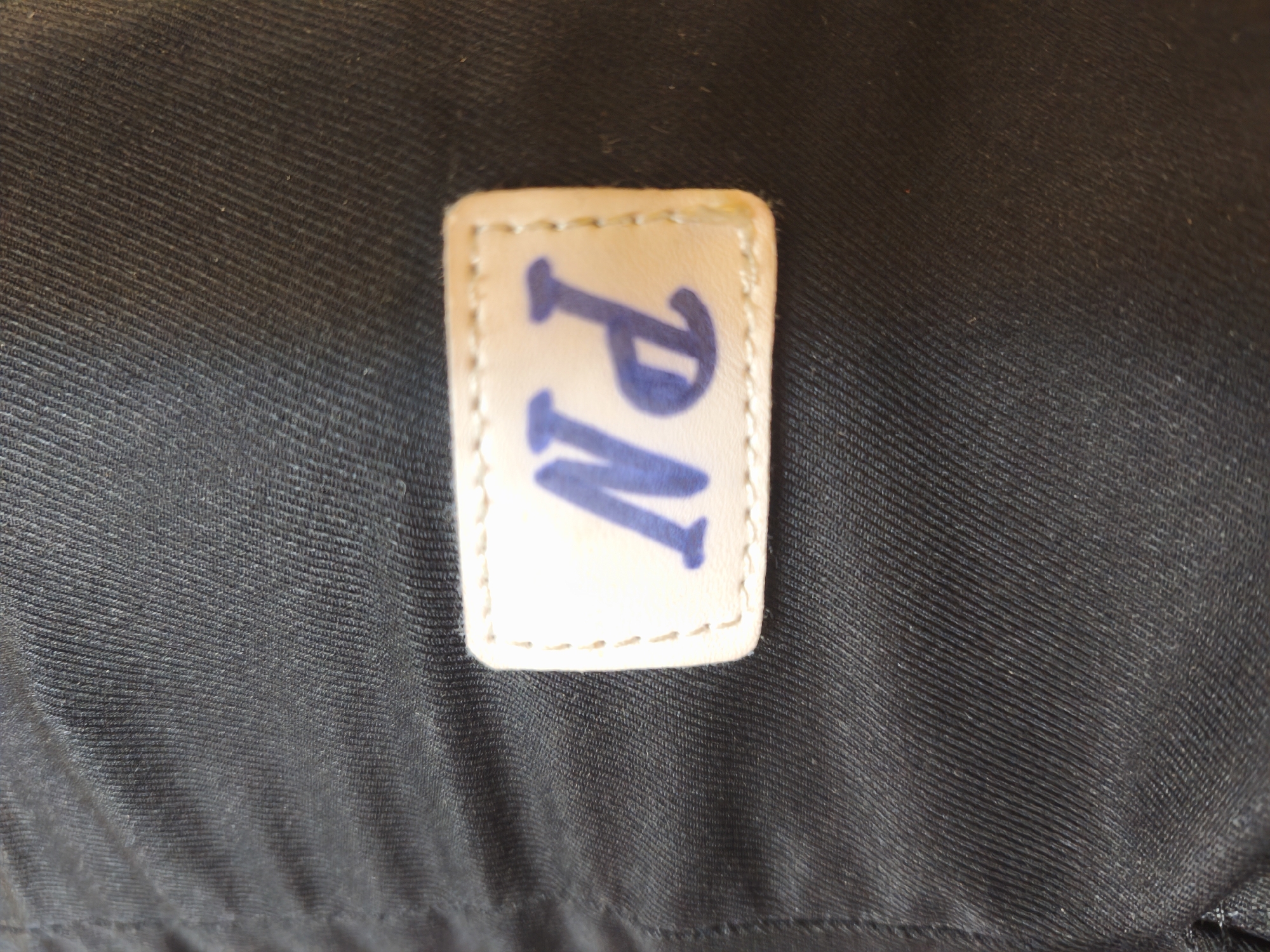 The image is a close-up shot of a black fabric with a small, rectangular, light-colored tag stitched onto it. The tag has the blue letters "PN" embroidered on it. The fabric appears to be textured, with visible horizontal lines. The lighting suggests an indoor setting, possibly a room in Kyiv, Ukraine, but no specific city landmarks or identifying features are present. No people or actions are depicted.