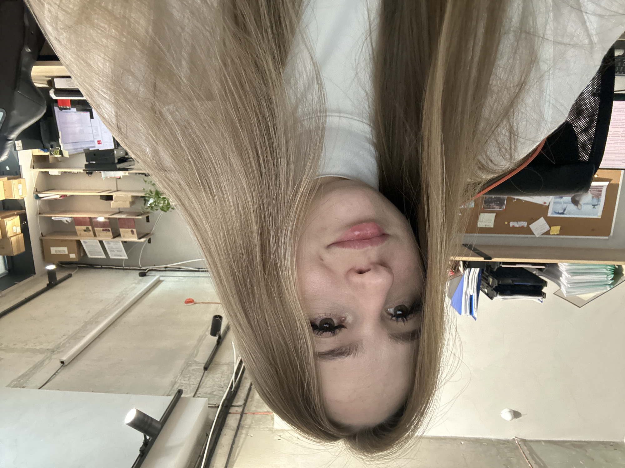 An indoor, eye-level, medium shot shows a young woman with long, blonde hair covering the right side of her face as she looks directly at the camera with a slight smile. She is wearing a white, high-necked sweater. The background appears to be an office environment. To the left, a shelf is stacked with papers and folders. In the mid-ground, a corkboard is visible with various notes and pictures pinned to it. A black office chair with mesh backing is partially visible on the left. To the right, shelves are stocked with boxes and other office supplies. The ceiling features exposed concrete, lighting fixtures, and wiring.