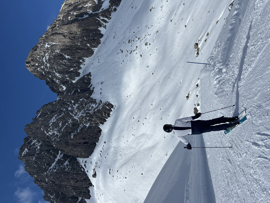 The image displays a solitary individual positioned on a snow-covered ski slope, facing away from the viewer towards a mountainous backdrop in the city of Unknown, Unknown. The person is equipped with a black helmet, a white long-sleeved top with a black vertical stripe on the back, dark ski pants, and black gloves, holding two ski poles. Their light blue-green skis with black bindings are parallel on the groomed snow. The background features two prominent, jagged, dark grey rock mountains, partially covered with snow, beneath a clear, deep blue sky. The snow-covered terrain exhibits visible ski tracks and scattered rocks on the upper slopes. Sunlight illuminates the scene from the left, casting distinct shadows from the skier and a small snow mound in the foreground, where two blue marker poles are present on the right side of the frame.