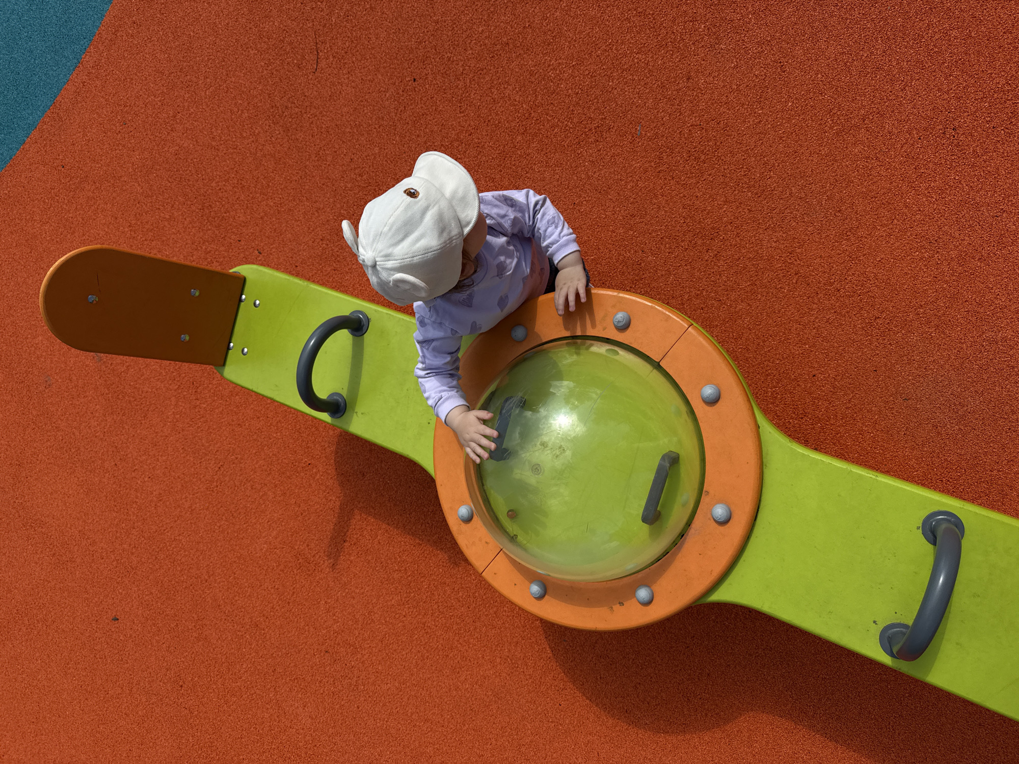A high-angle shot shows a toddler on a playground ride in Moscow, Russia. The toddler, wearing a white hat and a light-colored, long-sleeved shirt with a heart pattern, stands over a green and orange playground apparatus, touching the clear, green dome-like central part of the ride. The ride, which resembles a seesaw, has two green arms with dark grey curved handles at each end. The base of the playground is a textured surface, primarily orange, with a patch of blue in the upper left corner. Shadows cast by the ride and the toddler suggest a sunny day. The scene gives the impression of a typical, sunny day at a playground.