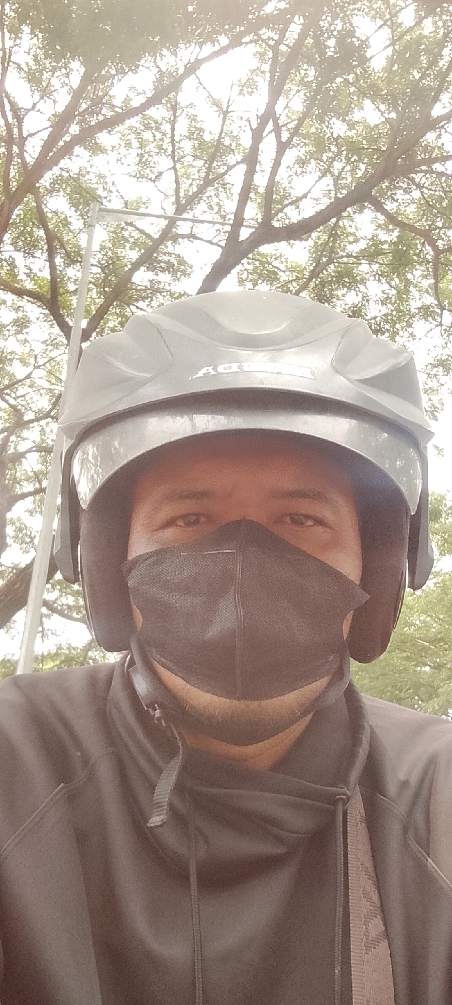 A medium shot, waist-up selfie of a person wearing a black face mask and a grey helmet. The person is outdoors, with a tree and sky in the background. The lighting is bright, suggesting daytime. The person is wearing a black long-sleeved shirt. The background shows the branches of a large tree and a bright sky. There is a vertical pole with a light fixture visible to the left of the person. The image appears to have been taken in Bekasi, Indonesia, given the surrounding foliage and the presence of a typical street light.