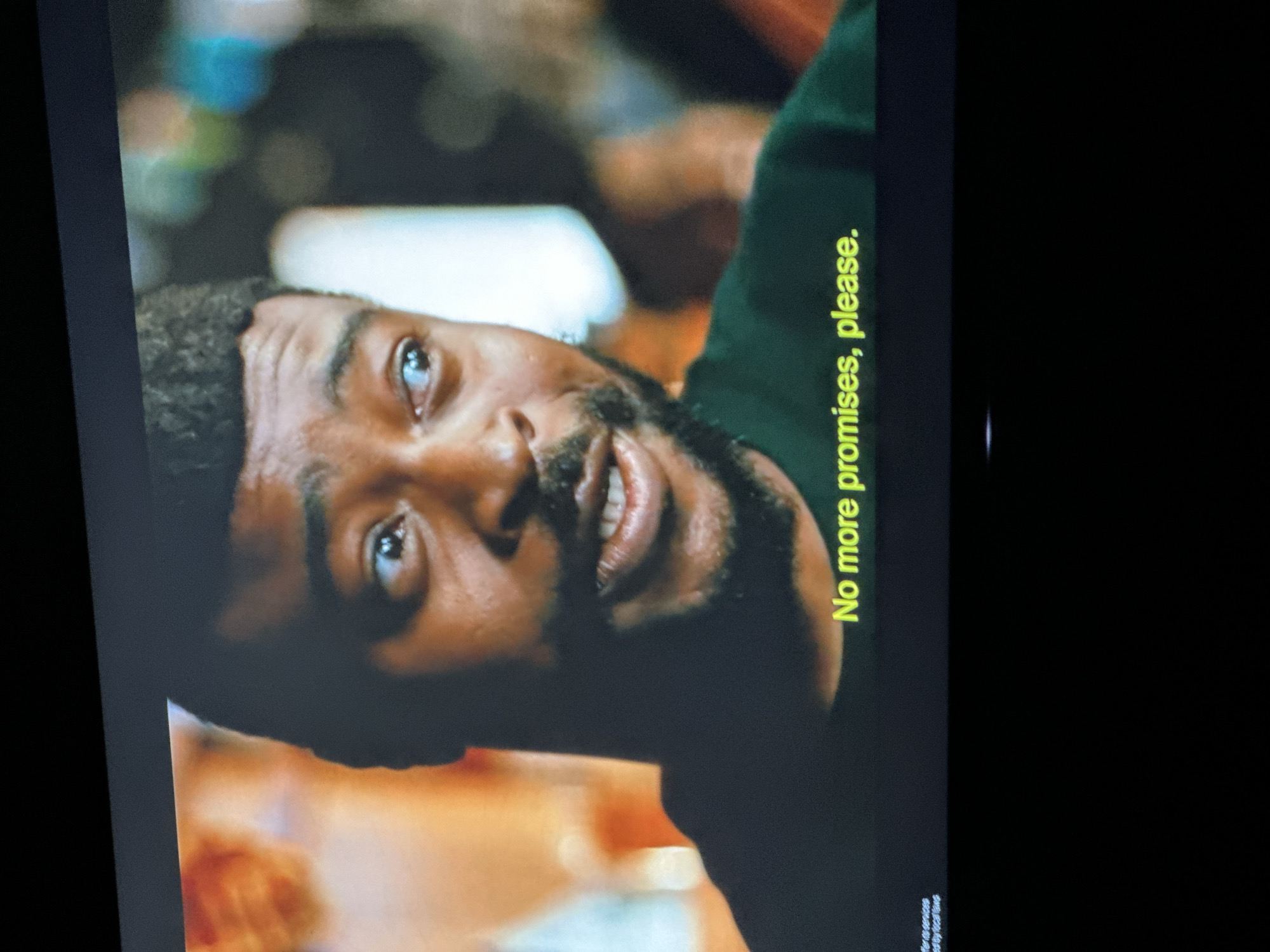 The image is a close-up shot of a Black man's face, with subtitles at the bottom reading "No more promises, please." The man has a short, dark hairstyle, dark eyes, and a light beard. He is wearing a dark green t-shirt. The background is blurred, with some indistinct lights and shapes, suggesting an indoor setting, possibly a bar or a social gathering. There is a small white text at the bottom left that is mostly illegible. There is no information in the image that could indicate the location in the city of Gura Top, Nigeria.