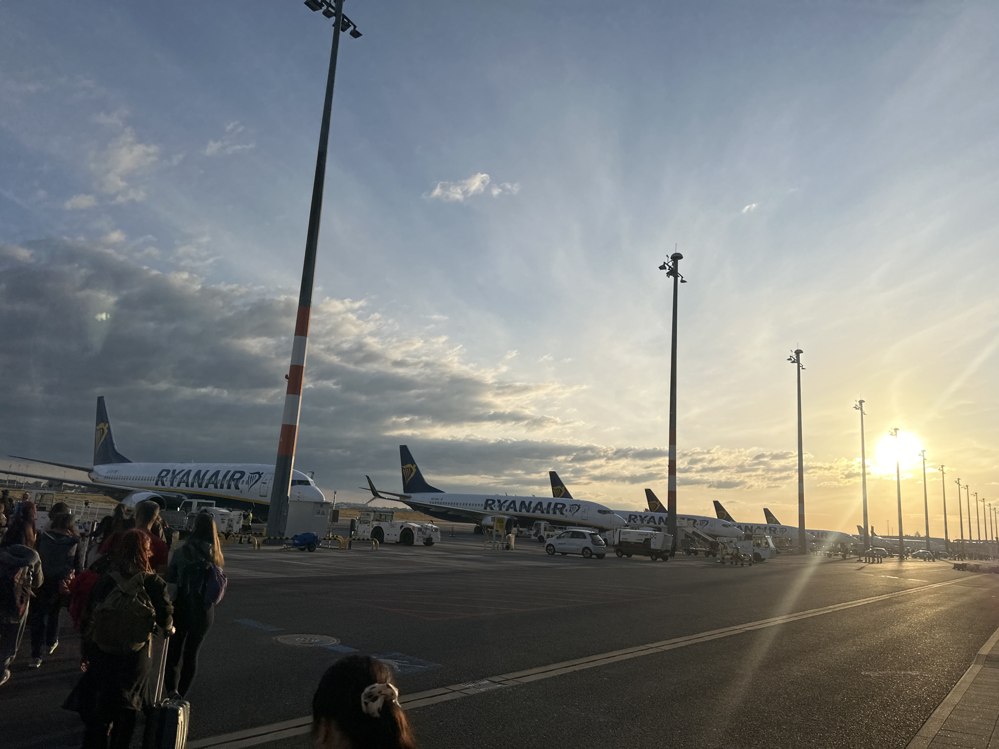 The image depicts an airport tarmac at sunset in Schönefeld, Germany. Several Ryanair airplanes are parked on the tarmac, with one prominently featured in the foreground, bearing the airline's branding. A group of people, appearing to be passengers, are visible standing in a queue, some with luggage, suggesting they are either boarding or disembarking from an aircraft. Tall light poles with multiple lamps are scattered across the scene, illuminating the area. The sky is partly cloudy, with the sun setting in the distance, casting a warm, golden light. The overall atmosphere is that of an active airport during a late hour.