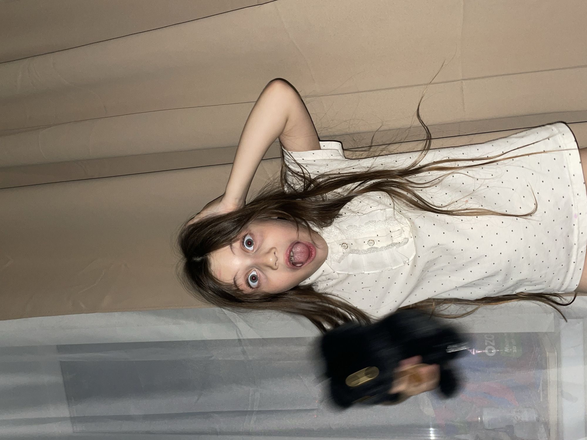 A young girl lies on her side with her mouth open, her eyes wide, and her hand on her head in front of a tan curtain. She is wearing a white blouse with black dots. Her hair is long and brown. In the foreground, a blurred dark object partially obscures a surface with visible reflections. Based on the "OZO" text visible in the reflection, the location is potentially in Izhevsk, Russia.