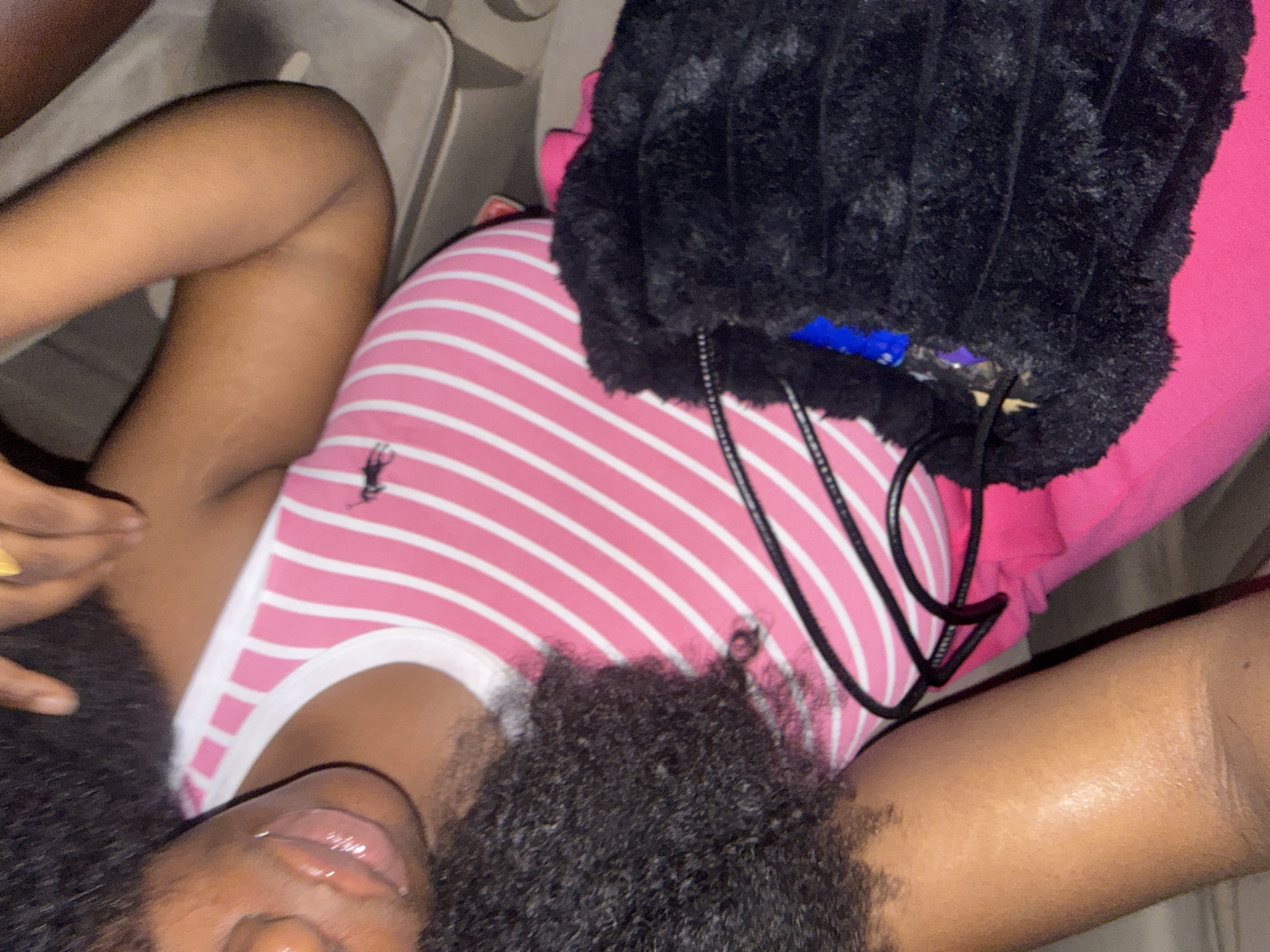 A person is lying down, wearing a pink and white striped tank top, with a black fuzzy bag next to them. The location is in what appears to be the passenger seat of a car. The city is Abuja, Nigeria. The person has dark skin and dark, curly hair. Their lips are glossy. The bag has drawstring cords. The interior of the car is visible.