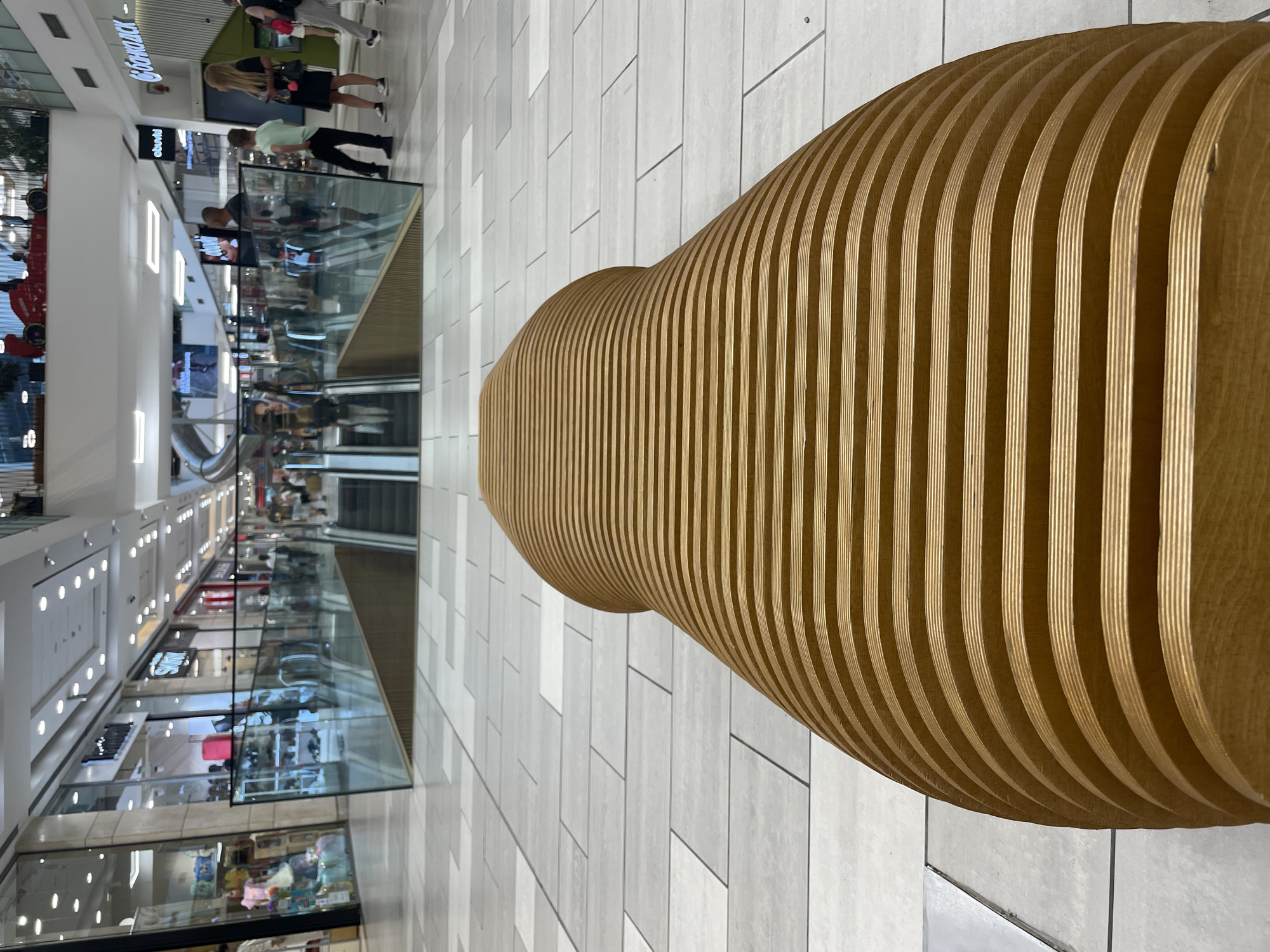 This image provides an interior view of Paradise Center, a shopping mall located in Sofia, Bulgaria.
In the immediate foreground, a large, curvilinear wooden bench or sculptural seating element dominates the lower half of the frame. It is constructed from numerous horizontally layered, light-brown plywood sheets, creating a ribbed, undulating surface that tapers towards the background. The camera is positioned low, looking over this distinctive piece of furniture. The floor beneath and around the bench is composed of rectangular tiles in varying shades of light grey and off-white, arranged in a staggered pattern with dark grout lines.
Beyond the foreground bench, a modern glass and light-brown wooden barrier extends across the midground, serving as a railing for a lower level. Behind this barrier, a set of descending escalators is visible, with several individuals on them. The glass reflects parts of the mall's interior, including overhead lights and people.
The background reveals multiple levels of the brightly lit shopping mall, lined with various retail storefronts. On the left side, through large glass windows, merchandise like children's clothing and shoes is displayed, with partial signage including "STORY" visible. On the right side, storefronts bear partially legible signs such as "abunti" and "ОББАНК" (UBBANK, a Bulgarian bank).
Numerous individuals are dispersed throughout the mall. In the mid-right background, a woman with long blonde hair wearing a black dress and white sneakers stands next to a young man in a light green t-shirt and dark pants. Further back, another person in grey pants with a backpack is seen. Other figures walk casually through the common areas and on the escalators.
Architectural and decorative elements are prominent on the upper levels. A large, red, sculptural object resembling a Formula 1 race car is suspended or displayed high above the right side of the mall's main thoroughfare. To the upper left, a large, curving, silver slide extends downwards from an upper level, indicating a leisure or play area. The high ceiling is fitted with numerous recessed circular spotlights and linear strip lights, providing ample illumination. The overall environment is modern, spacious, and appears to be a busy commercial and entertainment complex.