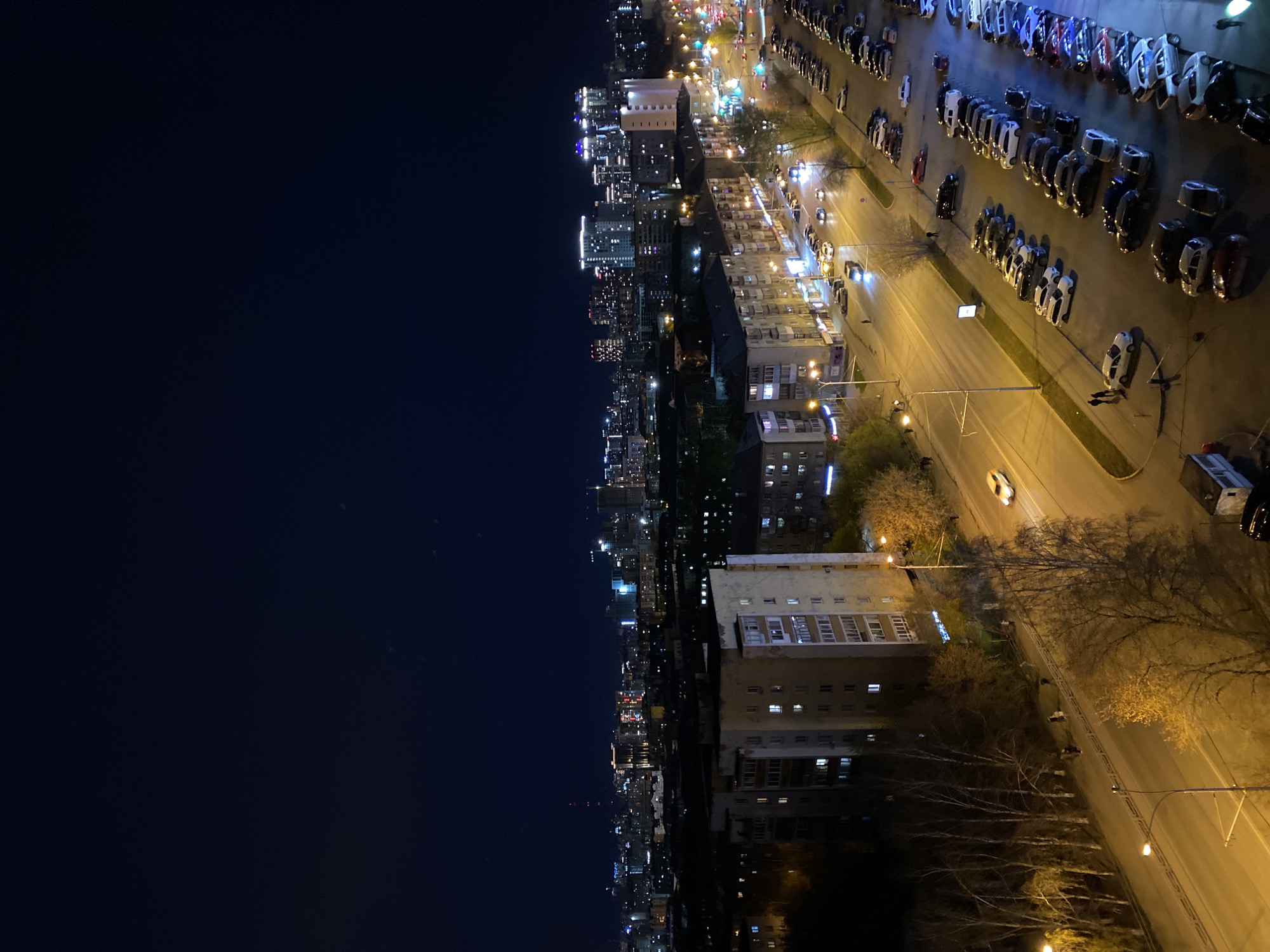 This high-angle, wide shot captures a city at night, presenting an expansive urban landscape illuminated by artificial lights. The vantage point is from an elevated position, likely a tall building.
On the right side of the frame, a multi-lane street stretches across the foreground and midground, brightly lit by numerous streetlights that cast a warm, yellowish glow. Parallel parking lots flank the street, filled with many parked cars, some reflecting the overhead lights. A white car is visible driving on the street, and a red car is parked on a side road. Towards the bottom right, a police vehicle with white and blue markings is partially seen. Bare trees, likely in late autumn or early spring, line parts of the street and stand between buildings. Several multi-story buildings, characterized by numerous windows and balconies, are prominent on this side, with many interior lights illuminating them.
The left side of the image reveals a sprawling cityscape extending into the distance. This area is a dense array of glowing lights from countless buildings of various heights, forming a vibrant and extensive urban skyline against the dark night sky. The sky itself is a deep blue-black, indicating a clear night.
The scene depicts typical urban activity after dark, with illuminated buildings suggesting ongoing life and movement of vehicles on the street. The overall impression is one of a vast, tranquil yet active metropolis. No people are visibly present, and no discernible text appears on any structures or vehicles. The context indicates this is a night scene in Yekaterinburg, Russia.