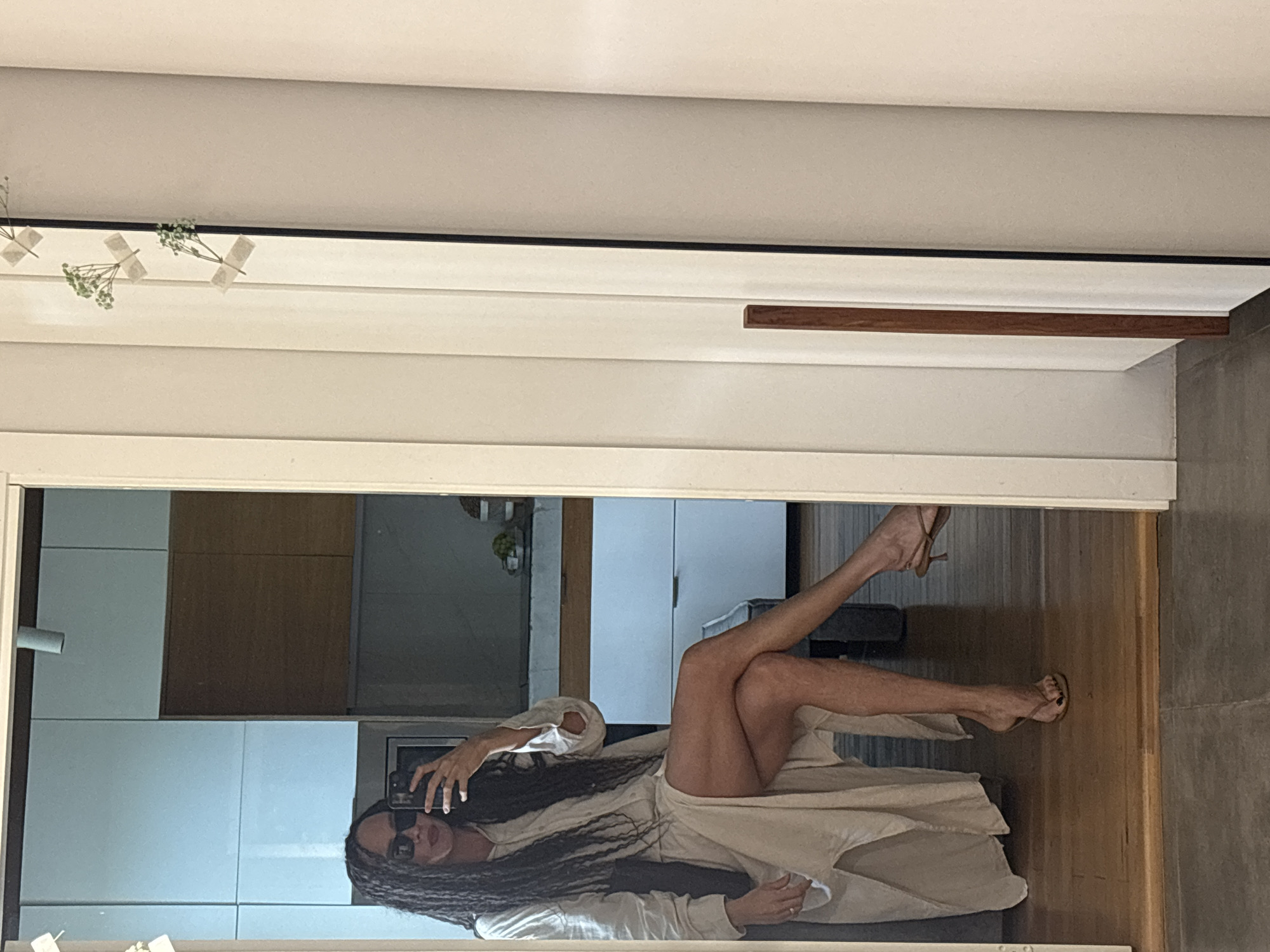 A woman is taking a photo of herself in a mirror. The woman is wearing sunglasses, a light-colored robe, and high-heeled sandals. She is lying on a bed or couch. The background appears to show part of an interior, possibly a kitchen and living area. There is no information to place this image in the city of Chaikovsky, Russia.