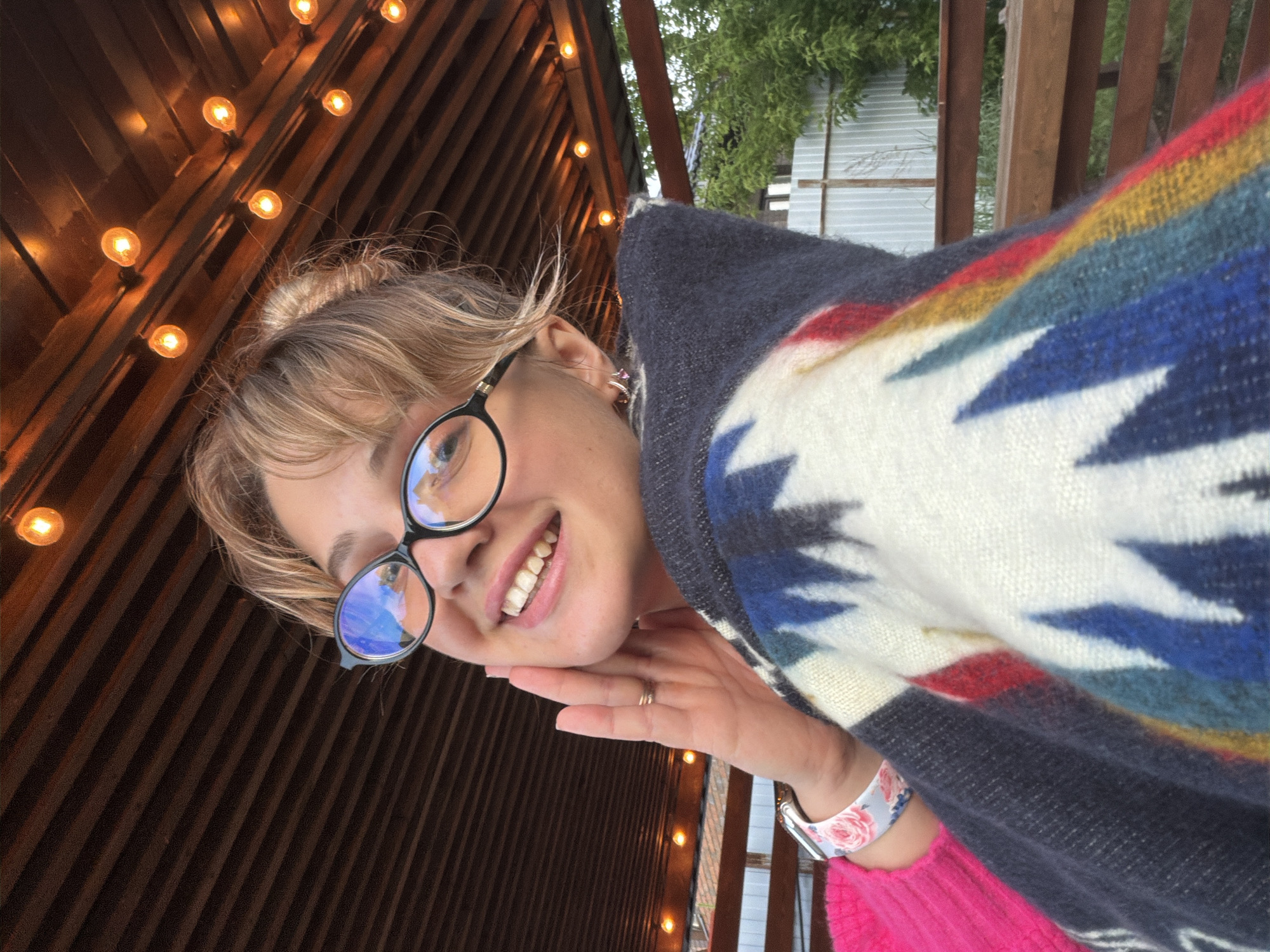 A young woman is captured in a tilted, close-up selfie, smiling broadly with teeth showing. She has blonde hair with bangs and appears to have some hair pulled back, with wisps around her face. She wears round, black-framed glasses that reflect a bright blue sky. Her left hand is positioned under her chin, and she wears a simple ring on her left ring finger. On her left wrist, a smartwatch with a floral-patterned band (white with pink flowers) is visible.
She is dressed in a thick, multi-colored poncho or blanket featuring a prominent geometric pattern in white, dark blue, light blue, red, and yellow, layered over a vibrant pink top or sweater.
The background suggests an outdoor setting, possibly a patio or balcony. It features dark wooden slats and beams, strung with warm, glowing Edison-style string lights. Beyond the wooden structure, green foliage from trees or bushes and a distant light-colored building are visible. The expression on the woman's face conveys happiness and contentment. No discernible text is visible in the image. The location context for the image is Большая Шильна, Russia.
