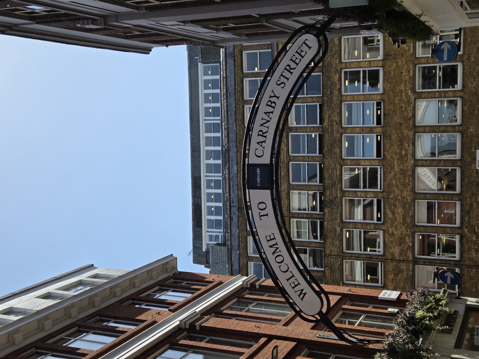 The image depicts a street sign that reads "Welcome to Carnaby Street." The street is located in the city of London, United Kingdom. The buildings in the background are a mix of brick and stone, and the sky is clear. A blue traffic sign showing direction is visible on the side of the building. Some greenery is visible alongside the walls of buildings.