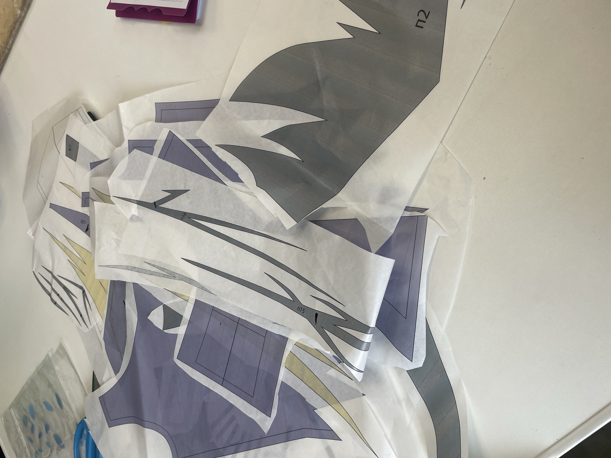 The image shows a pile of translucent pattern pieces, likely for sewing, spread across a white surface. The pattern pieces have lines and shaded areas in purple, grey, and yellow, and some are marked with letters and numbers like "n2," "C4," "C5," and "n15." In the background, there is a clear plastic bag with blue leaf-shaped cutouts and a purple binder. The scene appears to be indoors, possibly in a home or craft space, and is not located in Sinyavino, Russia.