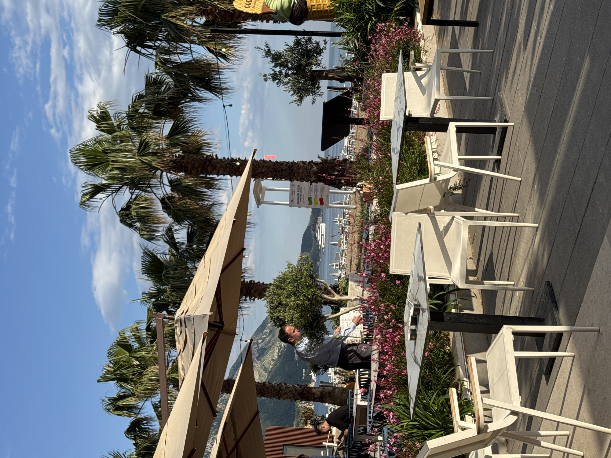 The image depicts an outdoor dining area in Rafailovići, Montenegro. Several white tables and chairs are arranged along a paved walkway. Palm trees and flowering plants provide shade and decoration. The background reveals a view of the coastline, with mountains and the Adriatic Sea visible. A man in a light-colored shirt and dark pants is visible near one of the tables. People are seated at some of the tables, while others stand nearby. Umbrellas provide shade over some tables. The sky is blue with scattered clouds.