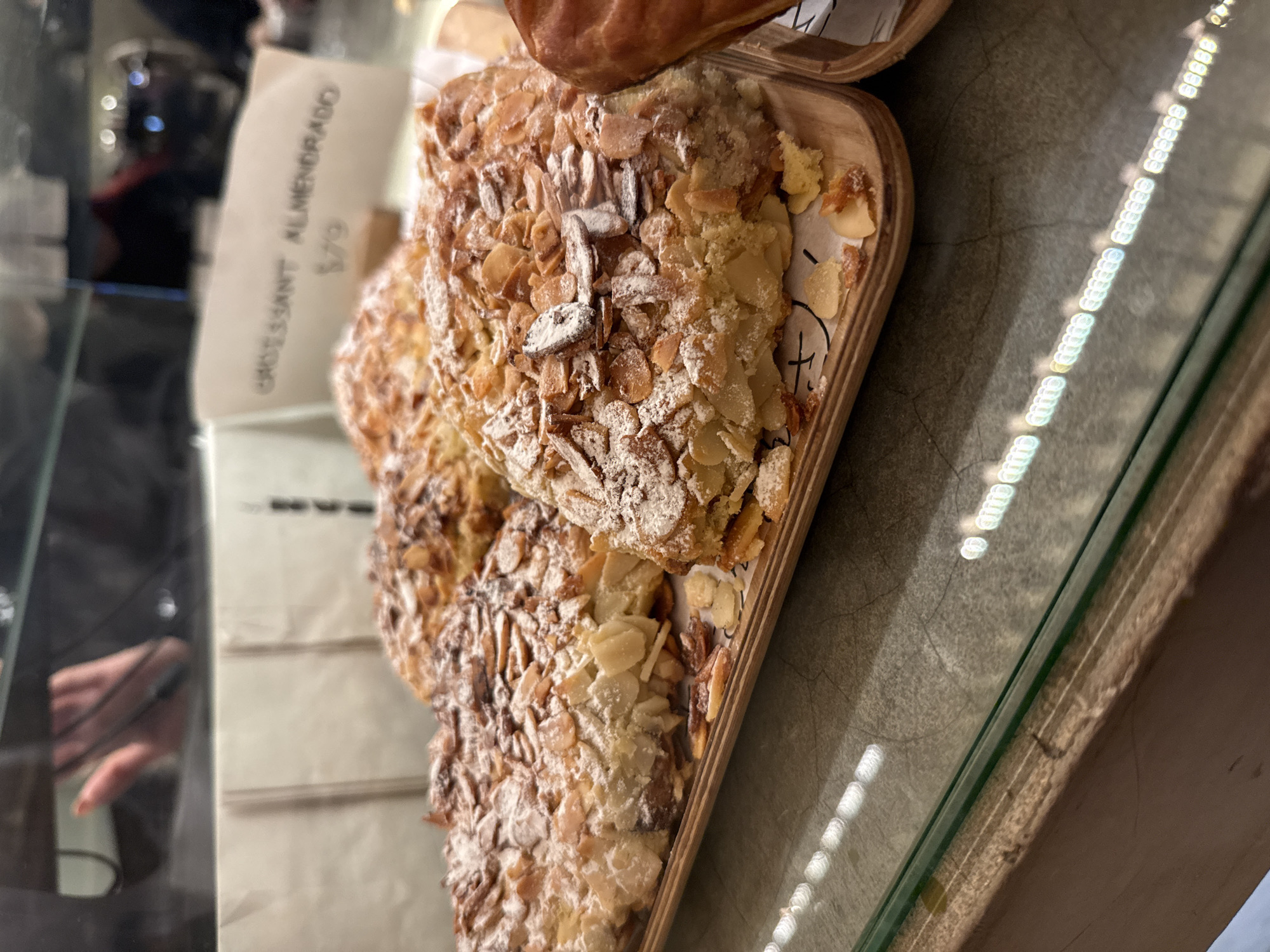 The image shows an almond croissant displayed in a wooden tray inside a glass display case. A label next to the croissant reads "Croissant Almendrado $79". The scene is likely a bakery or cafe in Mexico City, Mexico. Reflections in the glass display case show the blurred image of a person and other objects.