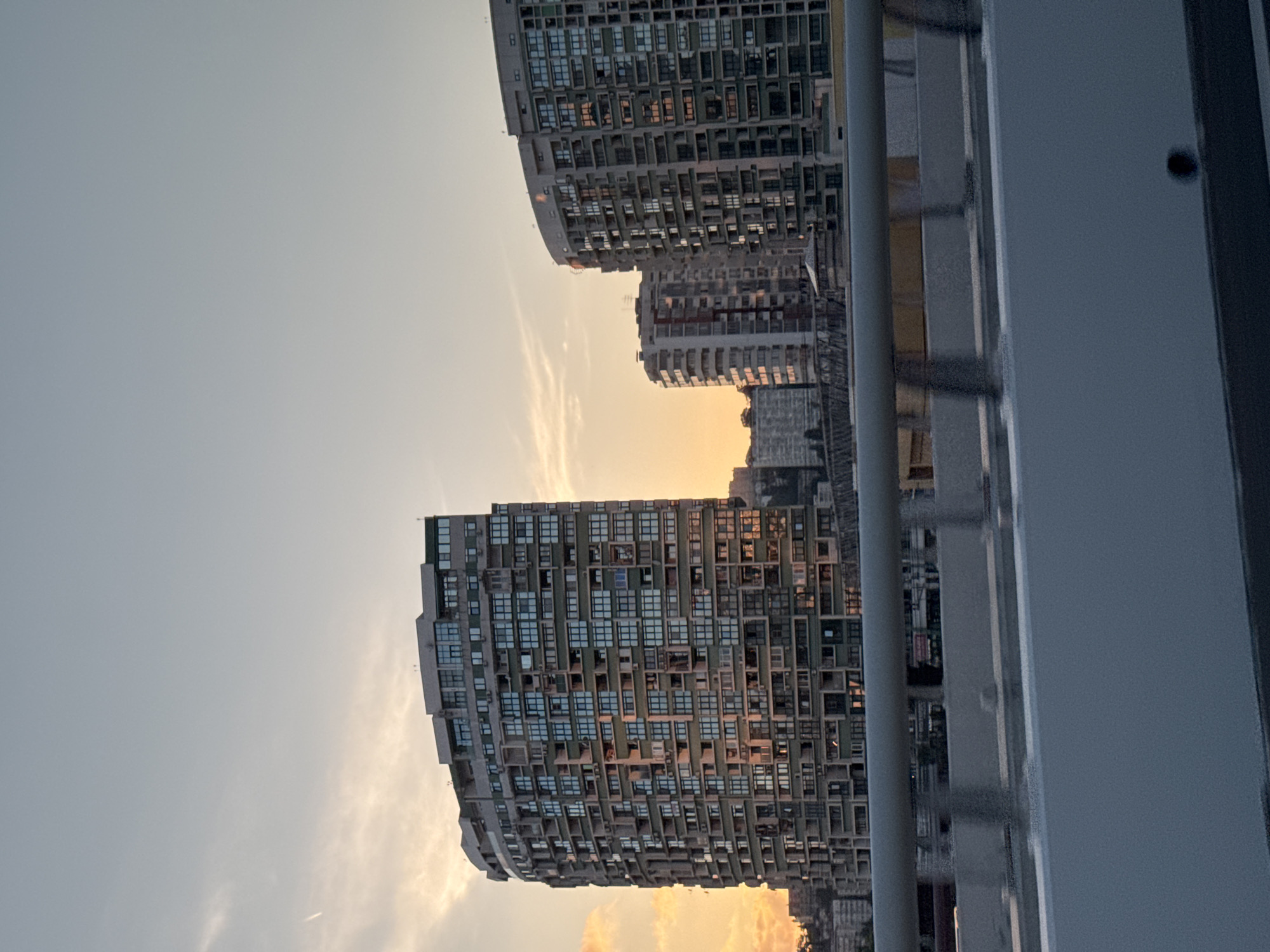 The image shows a cityscape with several apartment buildings at sunset. The sky is clear with some wispy clouds, and the setting sun casts a warm orange glow on the buildings. In the foreground, there is a bridge railing. The buildings are modern and feature many windows. The tallest building on the left has a curved facade and appears to be comprised of many individual balconies and window units. To the right, two other buildings are visible, one taller and more rectangular, and another shorter and also somewhat curved. The overall impression is one of urban development in a coastal area. This scene depicts a view of the buildings along the Sochi waterfront, likely taken from a bridge or elevated roadway, with the sun setting over the Black Sea.