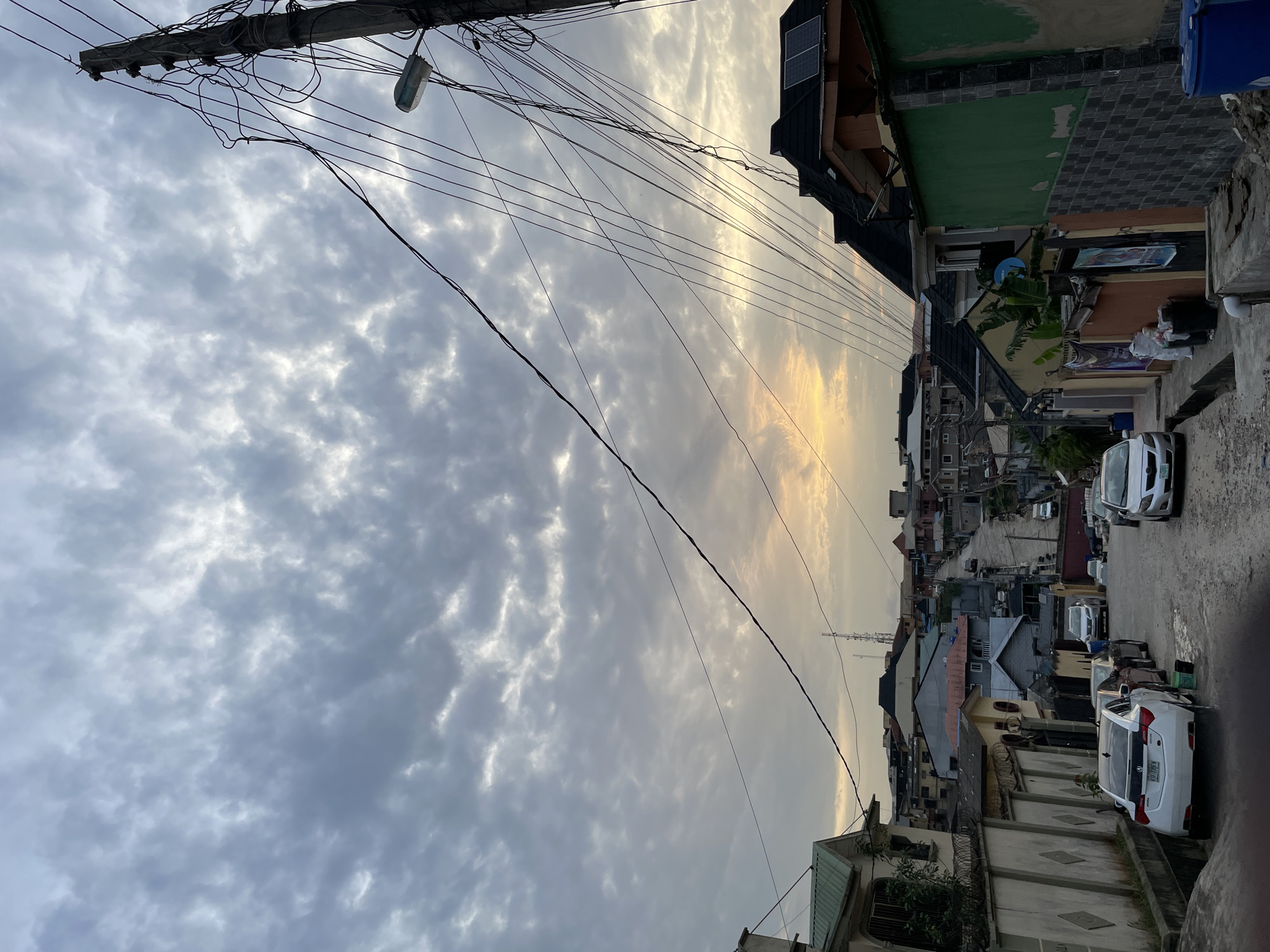 The image depicts a street scene in Isheri, Nigeria. Overcast skies with some golden light fill the majority of the frame. Utility poles and a dense network of electrical wires are visible at the top of the frame. The street is lined with buildings of varying heights and architectural styles, some featuring solar panels on their roofs. Parked cars line one side of the street. A person is walking on the right side of the road. The scene is taken from a low angle, looking upwards.