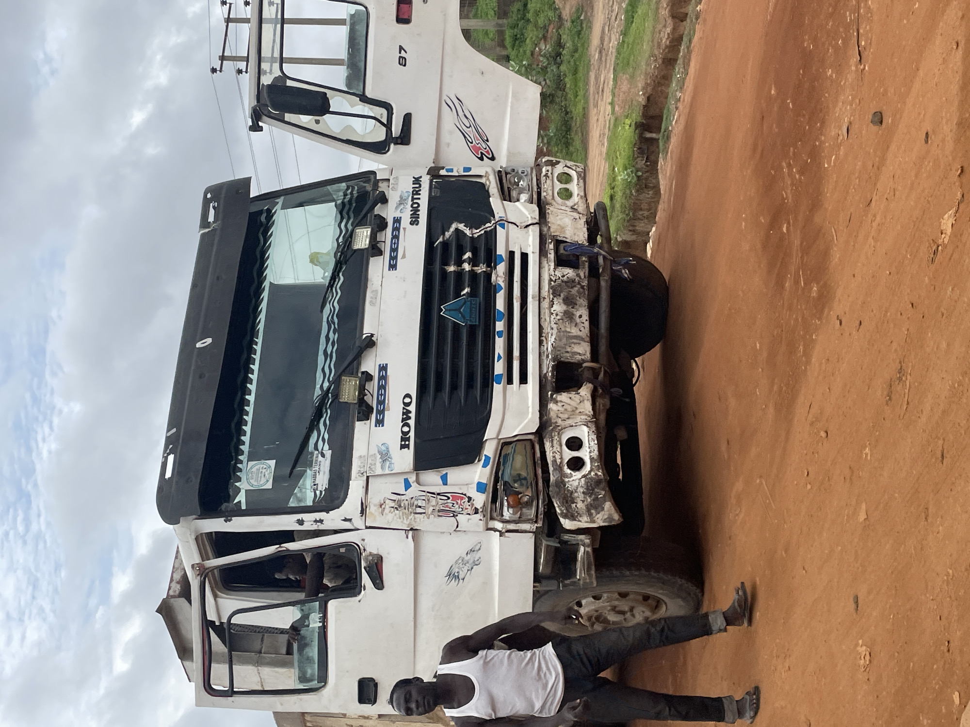A man in a white tank top and dark pants stands in front of a white Sinotruk truck on a dirt road. The truck is partially damaged, with a dented front bumper and a door ajar. The truck has the brands "HOWO" and "SINOTRUK" visible on its front. The man is looking away from the camera. The background shows a cloudy sky and some vegetation. The scene appears to be in Minna, Nigeria, as suggested by the context.