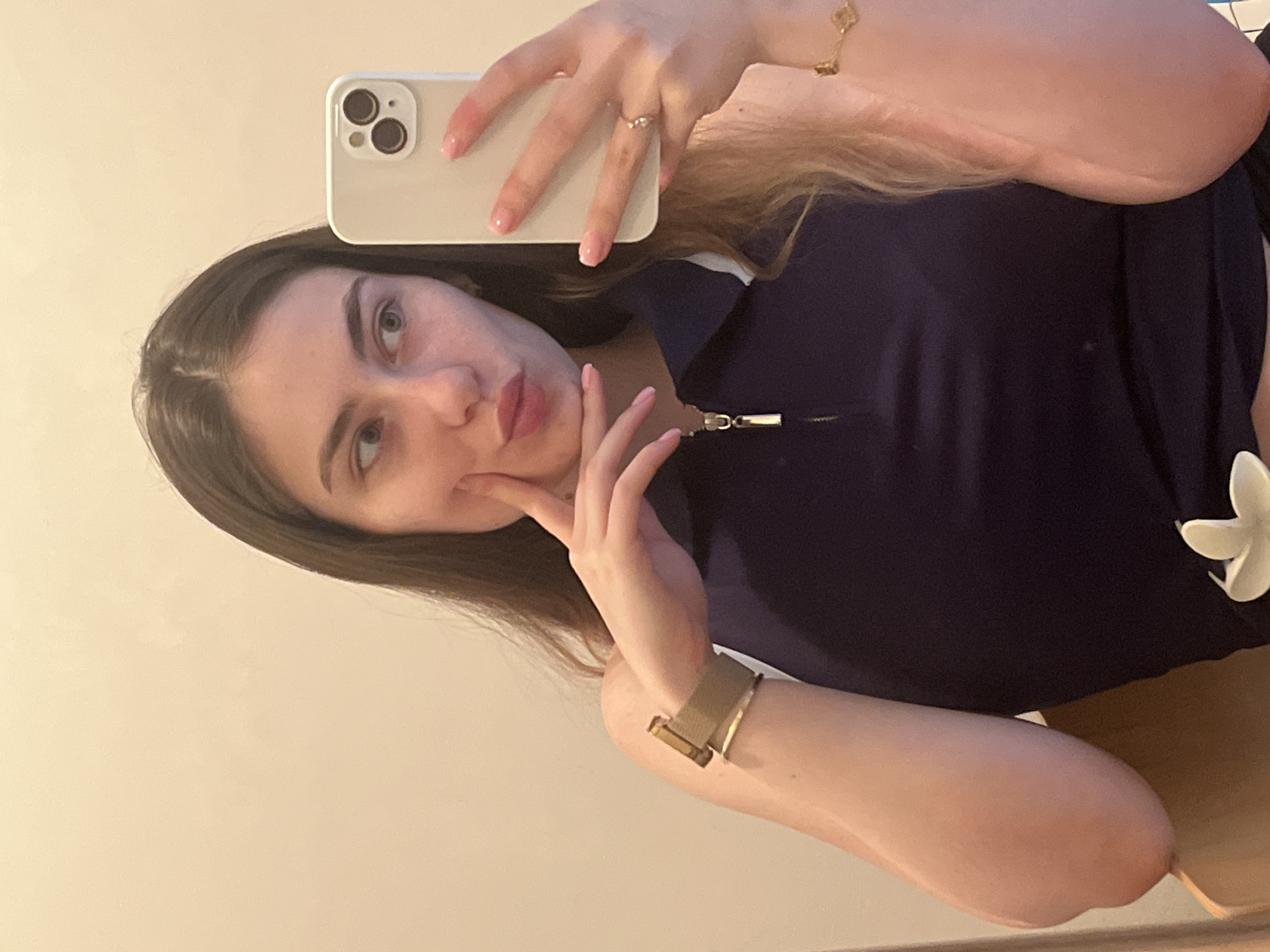 A mirror selfie of a young woman is captured indoors, presented with a 90-degree counter-clockwise rotation. The woman, with long, dark brown hair parted in the middle, light skin, and light-colored eyes, is visible from approximately the chest up. She wears a dark blue collared top with a zipper closure, under which a white shirt or trim is visible. A white, possibly floral, decorative element is attached to her top on the lower right side.
She holds a light-colored smartphone, likely an iPhone given its distinct camera module, in her right hand, positioned above her head. Her fingernails are manicured with light pink or nude polish. On her right wrist, she wears a gold-colored bracelet with clover-like charms, and a ring is visible on her right ring finger. Her left hand is posed near her chin/cheek, and a gold-toned watch with a mesh band is on her left wrist.
The woman looks towards the camera with a neutral expression, her lips slightly pouting. The background is a plain, light-colored wall, indicating an indoor setting. No specific location cues, time of day indicators, or visible text are present in the image.