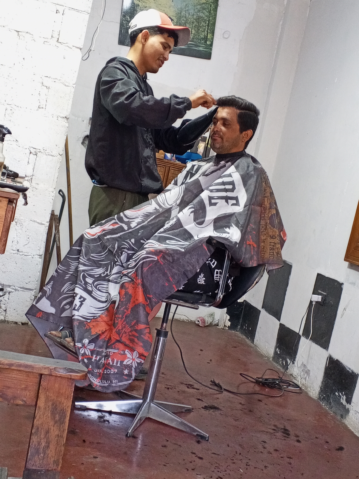 A medium shot shows a barber cutting the hair of a seated male customer. The barber, dressed in a black hoodie and cap, stands behind the customer and meticulously trims his hair. The customer, draped in a patterned cape, sits in a barber chair, his expression calm.
The setting appears to be a small barbershop, indicated by the barber's tools visible on a nearby table. The shop features a unique checkerboard pattern near the floor. A painting hangs on the wall behind the barber, and a power outlet is visible on the wall next to the customer's chair. Debris litters the floor around the chair, likely from the haircut. The text "AED HAALI JAN. 2007 HONOLULU, HI" is visible on the cape.