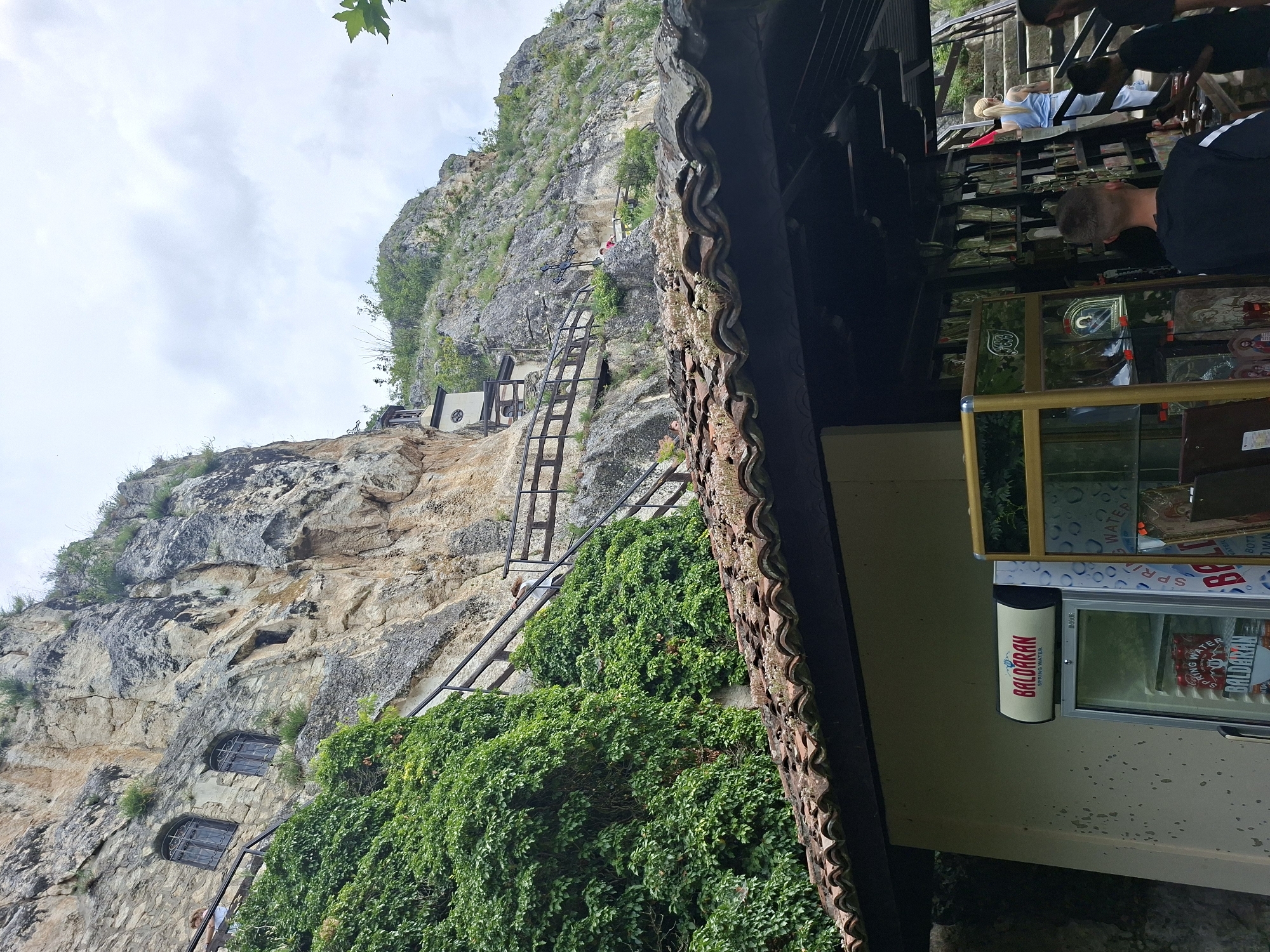 This image depicts the Basarbovo Rock Monastery in Basarbovo, Bulgaria. The primary subject is a rugged, light-colored rock cliff face that rises steeply, dominating the upper two-thirds of the frame. The cliff is partially covered with dense green foliage, particularly on the left side, and sparse vegetation on its upper ledges.
Integrated into the cliff on the left are two arched windows with dark, possibly wrought-iron grilles, set within a stone-built section. To their right and further up, a smaller rectangular opening or window is also present in the rock. Wooden staircases with dark railings ascend the cliff face, winding around its contours. Several individuals are visible on these stairs; one person with light hair is seen from behind, leaning on a railing on the upper left section of the stairs, and another person in a pink shirt is higher up on the right, near a large, dark crucifix mounted on a rocky ledge. Further up the cliff and to the right, a small, light-colored building with a dark roof and a small belfry (bell tower) is visible. The sky above is overcast with light grey clouds.
In the foreground, a terracotta-tiled roof extends across the middle of the image, covering a structure below. Beneath this roof, on the left, is a light-colored building wall. Against this wall stands an upright glass-front refrigerator labeled "BALDARAN SPRING WATER," containing visible cans of beverages. To the right of the refrigerator, under the same roof, is a display area, likely a kiosk or souvenir shop. It features multiple shelves and glass display cases containing what appear to be religious icons and other merchandise. Several individuals are present in this foreground area: a male in a black t-shirt with white stripes on the sleeves stands facing away from the camera, seemingly browsing the display. To his right, a female with blonde hair, wearing a light blue sleeveless top and light-colored pants, is walking up a set of stone stairs that ascend from the right side of the frame. Another person is partially visible on these stairs on the far right.