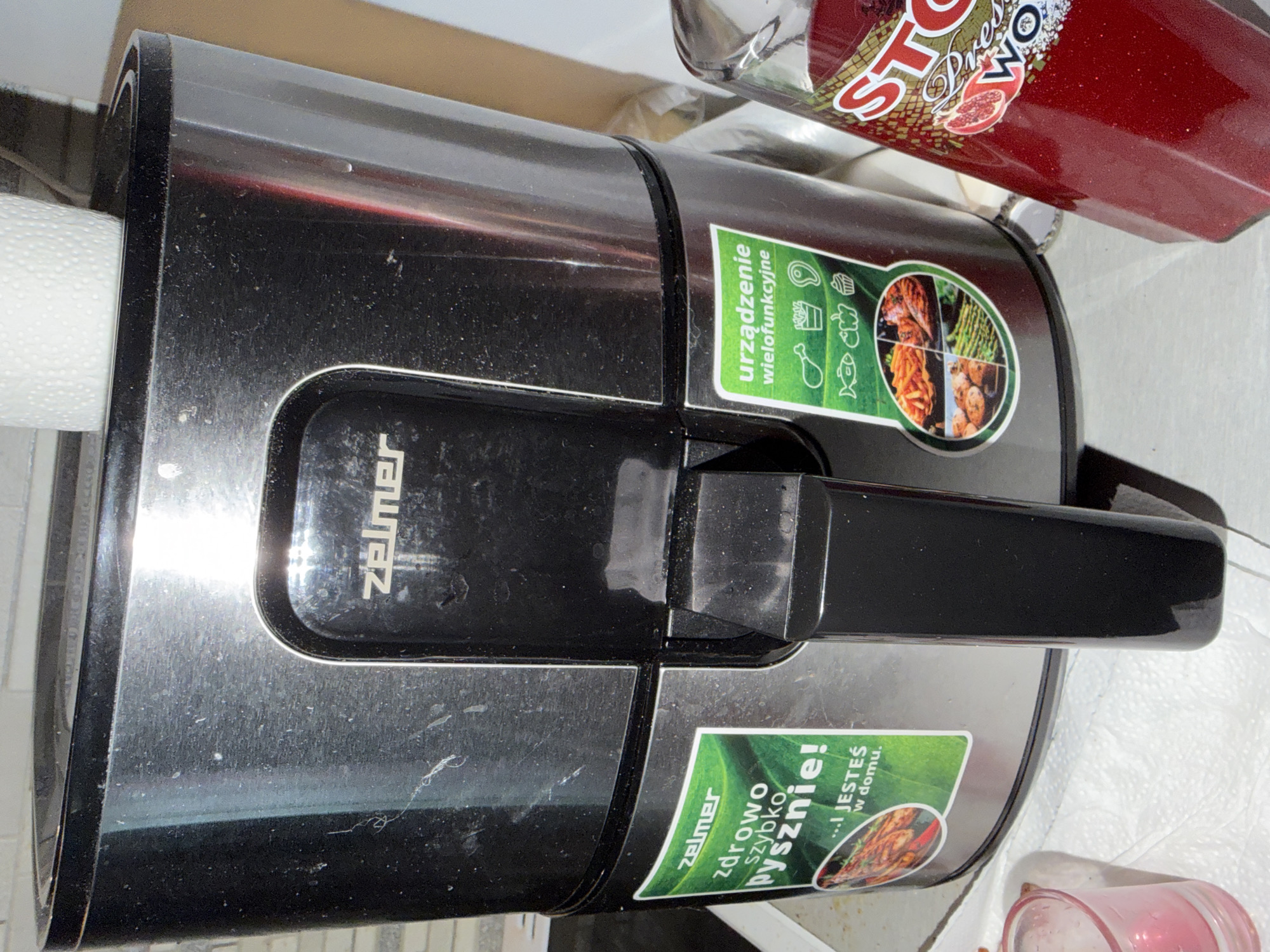 The image shows a Zelmer air fryer on a countertop. The air fryer is primarily black and silver, with a black handle. Two green stickers are visible on the side of the air fryer. The left sticker reads "zelmer zdrowo szybko pysznie! ...! JESTEŚ w domu." and features an image of grilled food. The right sticker reads "urządzenie wielofunkcyjne" and displays icons representing chicken, fries, steak, fish, cupcake, and other food items, along with a circular image divided into quadrants showing fries, grilled chicken, potatoes, and asparagus. A partially visible bottle of red juice with a label that says "STORM" is to the right of the air fryer. A white paper towel is draped over the top of the air fryer. A small pink glass is visible on the left side of the image. The overall environment appears to be a kitchen. The image does not contain any information that would allow for determination of its location in Szczecin, Poland.