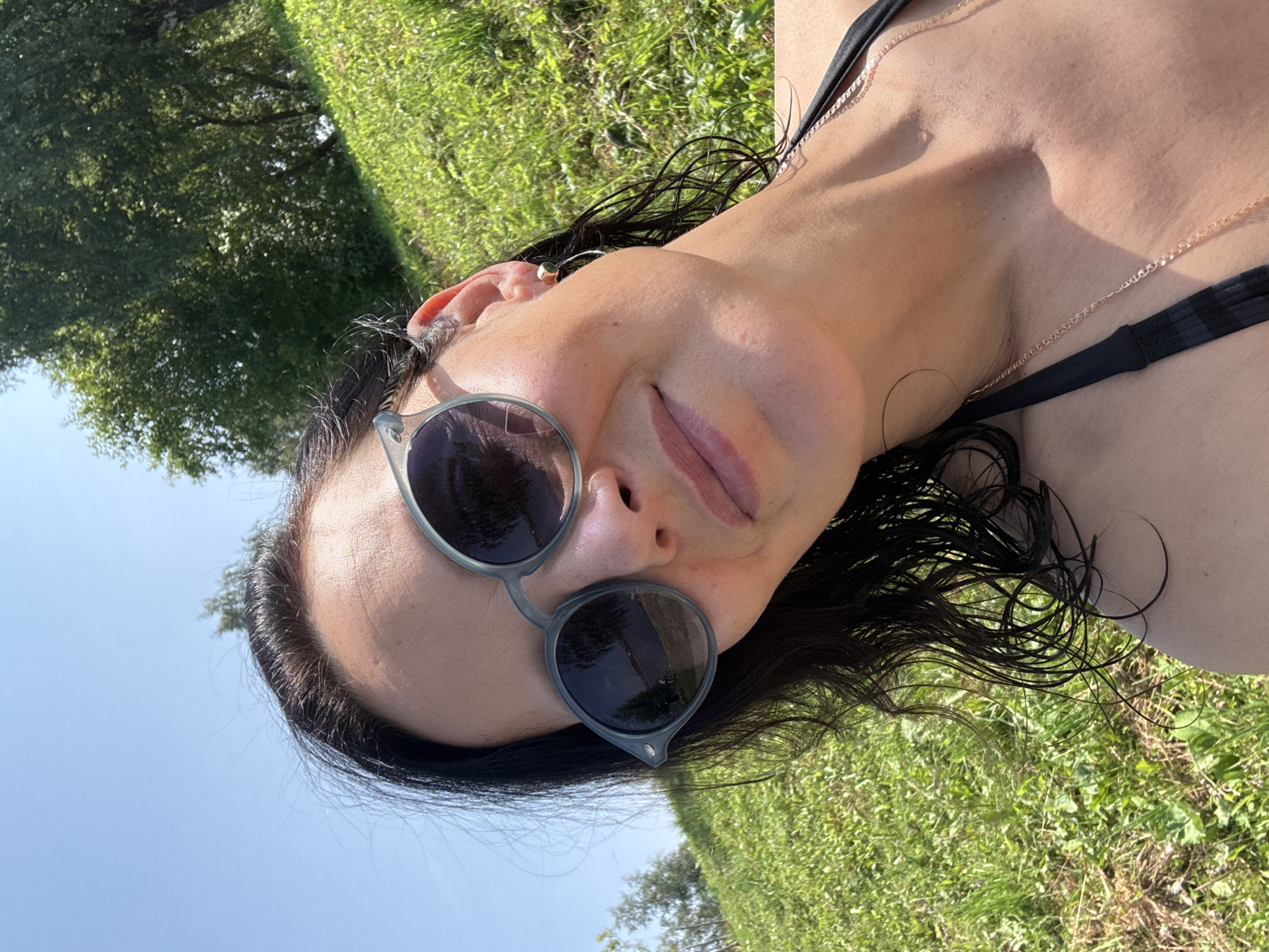 A woman is taking a selfie outdoors on a sunny day, possibly near a body of water, as indicated by the reflection in her sunglasses. She is wearing a black bikini top and sunglasses. Her dark hair is wet and falls around her face and shoulders. The background consists of lush green grass and trees, suggesting a natural, possibly park-like setting. The lighting suggests it is daytime. The location in Lysva, Russia, is not visually evident from the image itself.