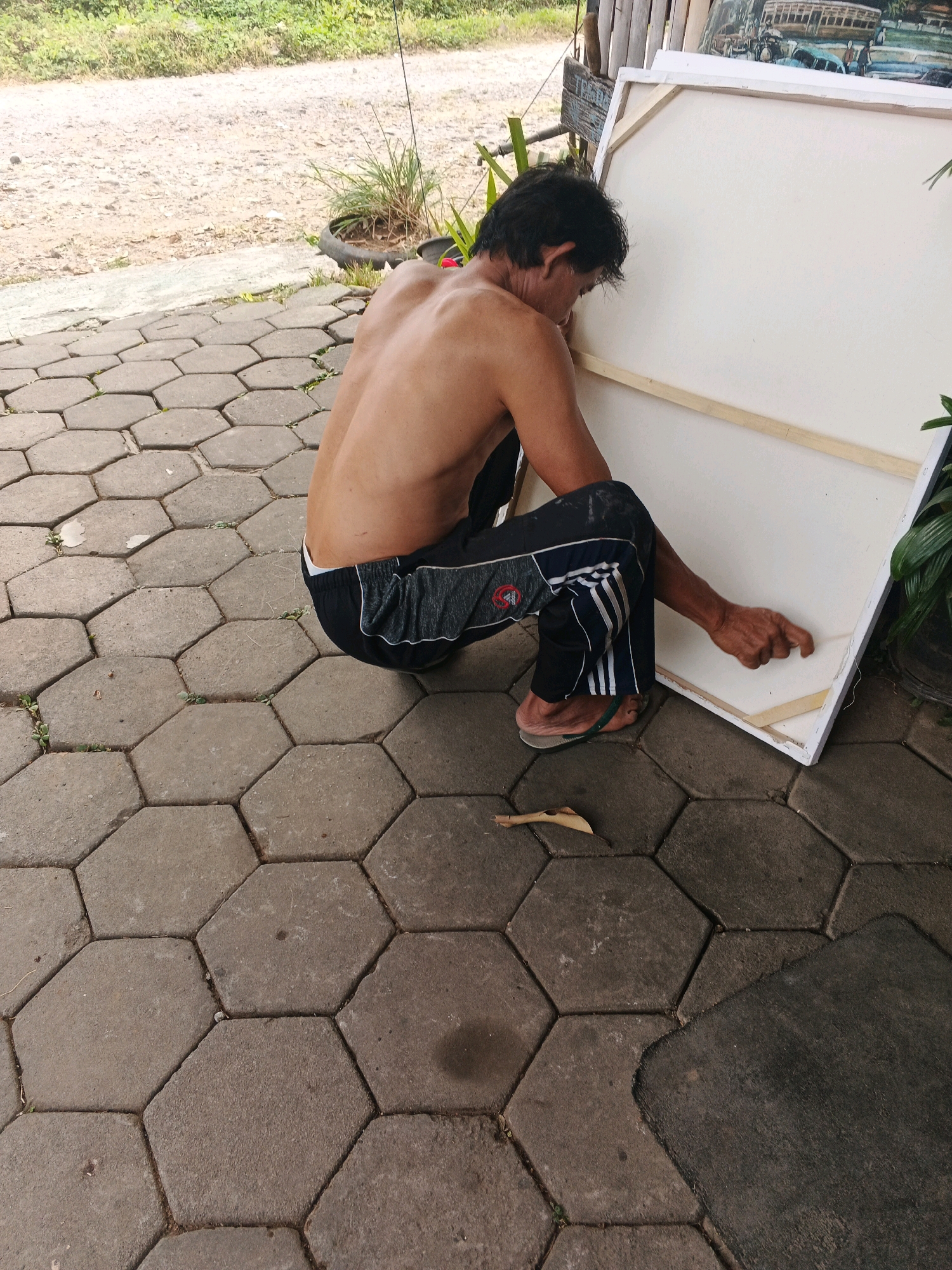 A shirtless person with dark hair is crouching on a hexagonal paver patio in front of a large white canvas propped on wooden supports. The person is wearing dark pants with white stripes and flip-flops. Their right hand is reaching towards the canvas. The background shows a dirt path and vegetation. The scene is outdoors, possibly in Ungaran, Indonesia.