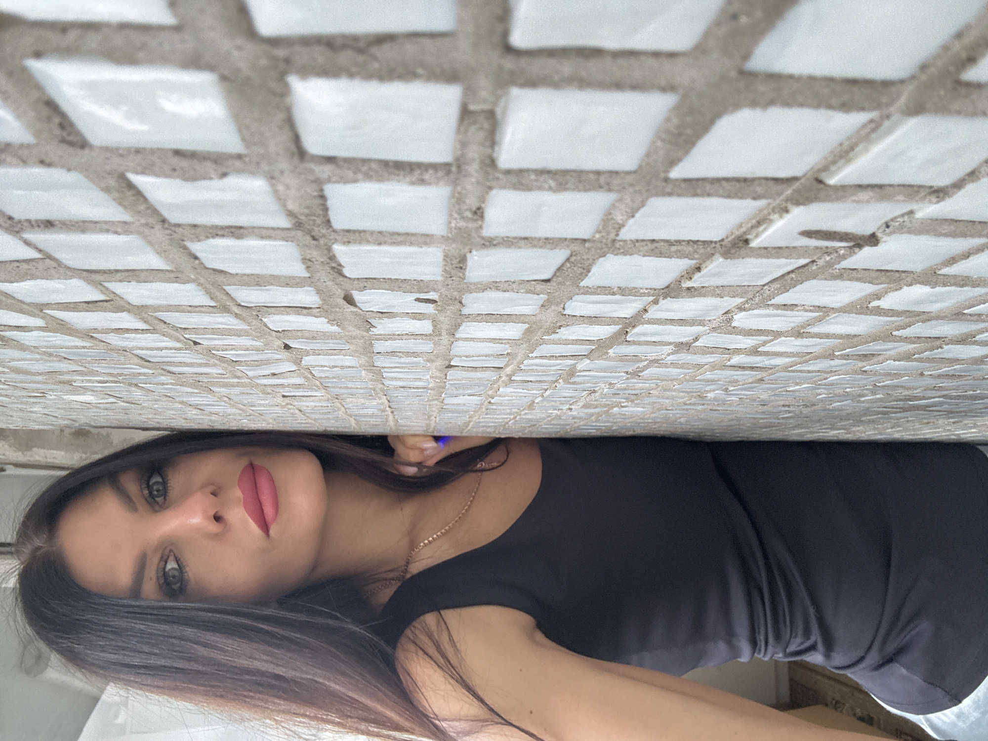 The image presents a portrait of a young woman taken from an unconventional, rotated perspective, where the subject appears to be lying down with her head near a tiled surface, or peeking from beneath it. When viewed upright, the woman occupies the lower portion of the frame, looking directly towards the camera.
The primary subject is a young woman with long, dark brown hair that falls over her shoulders and chest. She has light-colored eyes, possibly green or hazel, and is wearing a soft, muted pink or reddish lipstick. Her expression is neutral and direct. She is dressed in a plain, dark-colored top, likely black, and wears a delicate gold chain necklace around her neck.
The background is dominated by a wall or ceiling covered in small, off-white or light grey square tiles. These tiles are set with wide, dark grey grout lines, and their surfaces show some texture and slight irregularities, suggesting an older or rustic style. The tiled surface recedes into the background, creating a sense of perspective. The lighting is soft and diffused, indicating natural daylight, possibly indoors or under an overhang. There is no visible text in the image. The scene is set in Chaikovsky, Russia.