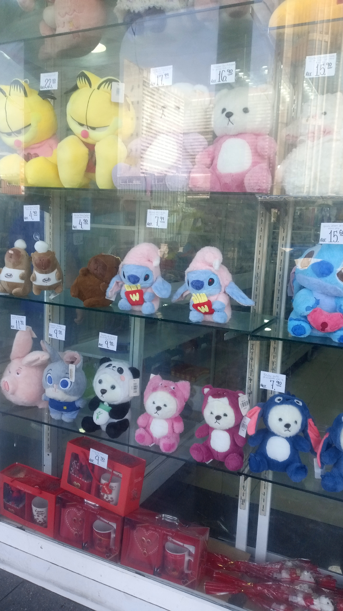The image depicts a display of plush toys in a glass-fronted cabinet or window, possibly in a store or retail setting. A variety of stuffed animals are visible, including depictions of Garfield, bears, and Stitch characters, each with price tags attached. In the foreground, below the toys, there are red gift boxes, some containing mugs. The window reflects an outside view.