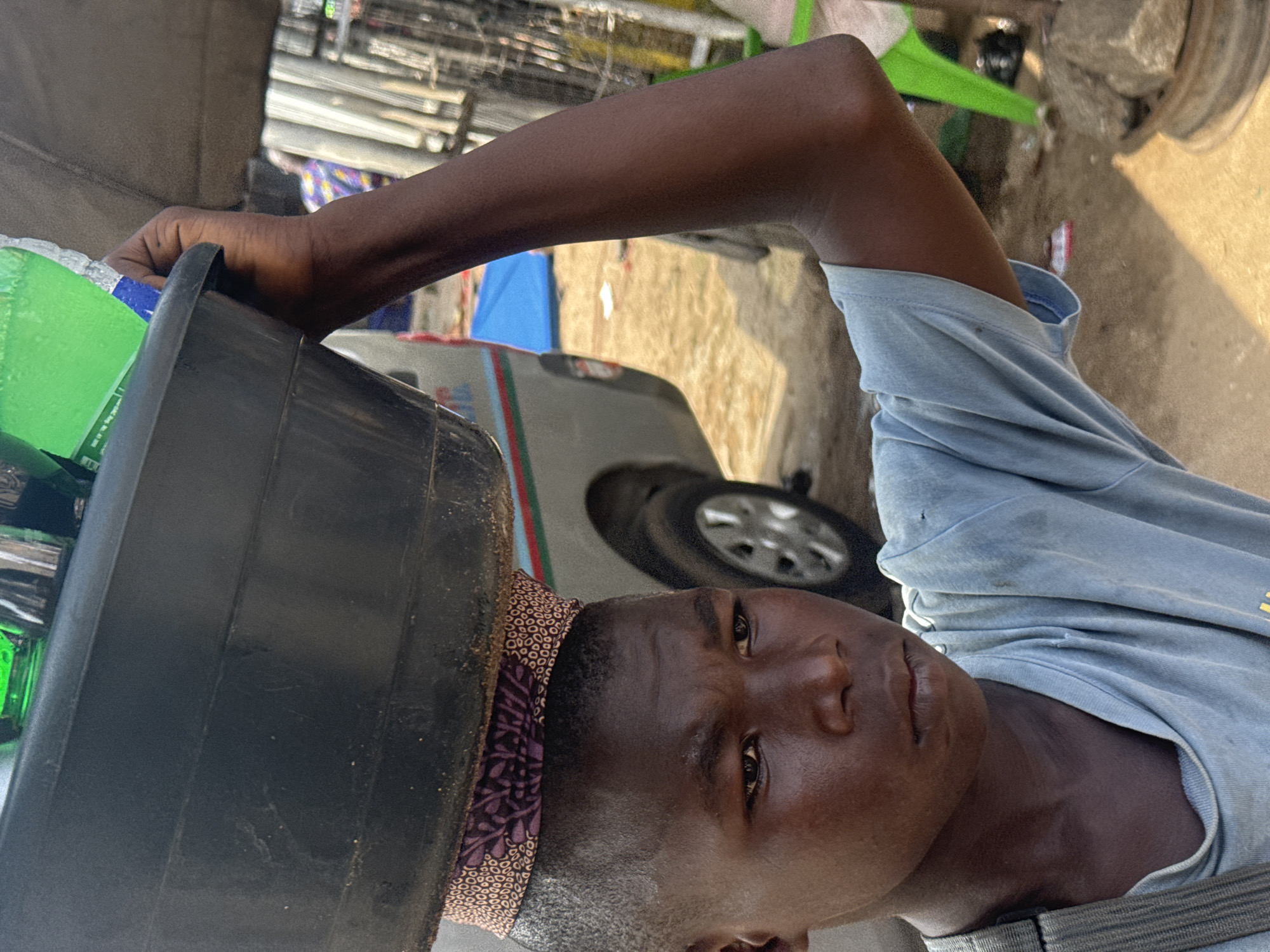 A young Black boy, appearing to be of school age, is seen from the chest up, carrying a large, black plastic basin filled with green plastic bottles on his head. The basin is balanced on a patterned fabric band around his head. He is wearing a light blue t-shirt, which is slightly worn and has some dirt stains. His expression is serious and he is looking slightly to his left. His right arm is raised to support the basin. The background appears to be an outdoor market setting in Nkpor, Nigeria. A silver van is partially visible behind him, and there are hints of market stalls, a blue tarp, and people in the distance. The ground is sandy and uneven.