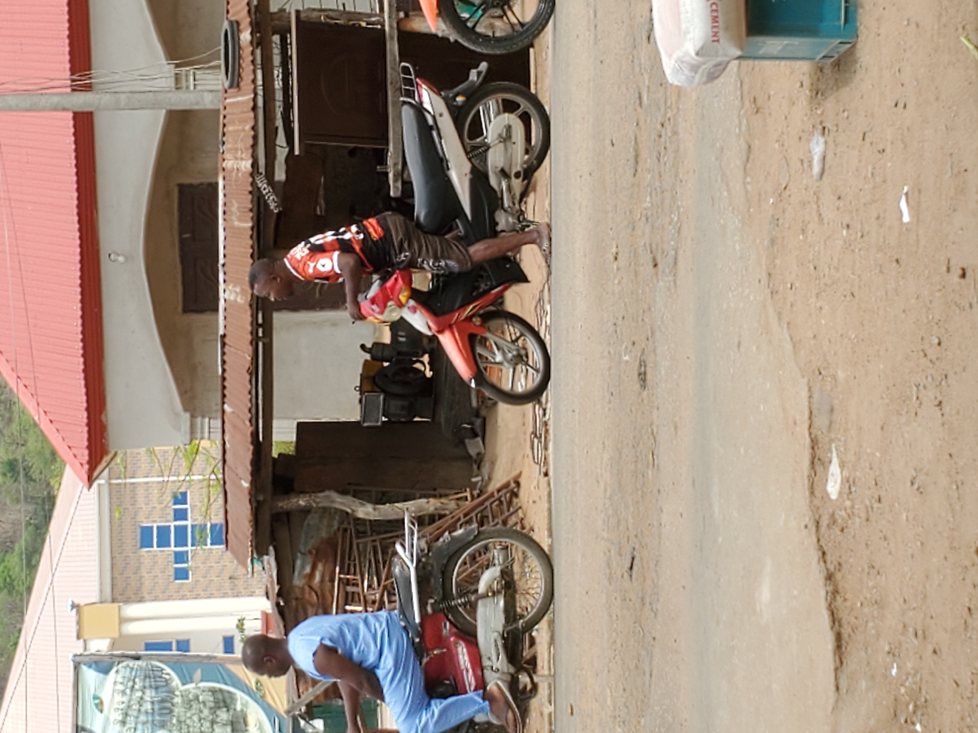 The scene is an outdoor shot, possibly in Mararaba, Nigeria, judging by the style of the buildings and the activities depicted. Two men are seen interacting with motorcycles. One man, wearing a blue outfit, is bending over a red motorcycle on the left side of the frame. Another man, wearing a black and orange top and brown shorts, is leaning over a red and grey motorcycle on the right side of the frame. There are at least three motorcycles visible, two in the foreground and another partially visible in the background. A building is behind the men, with a corrugated metal roof. The road surface is dirt. Cement bags are visible in the top right corner. Another building in the background has a cross design made of windows.