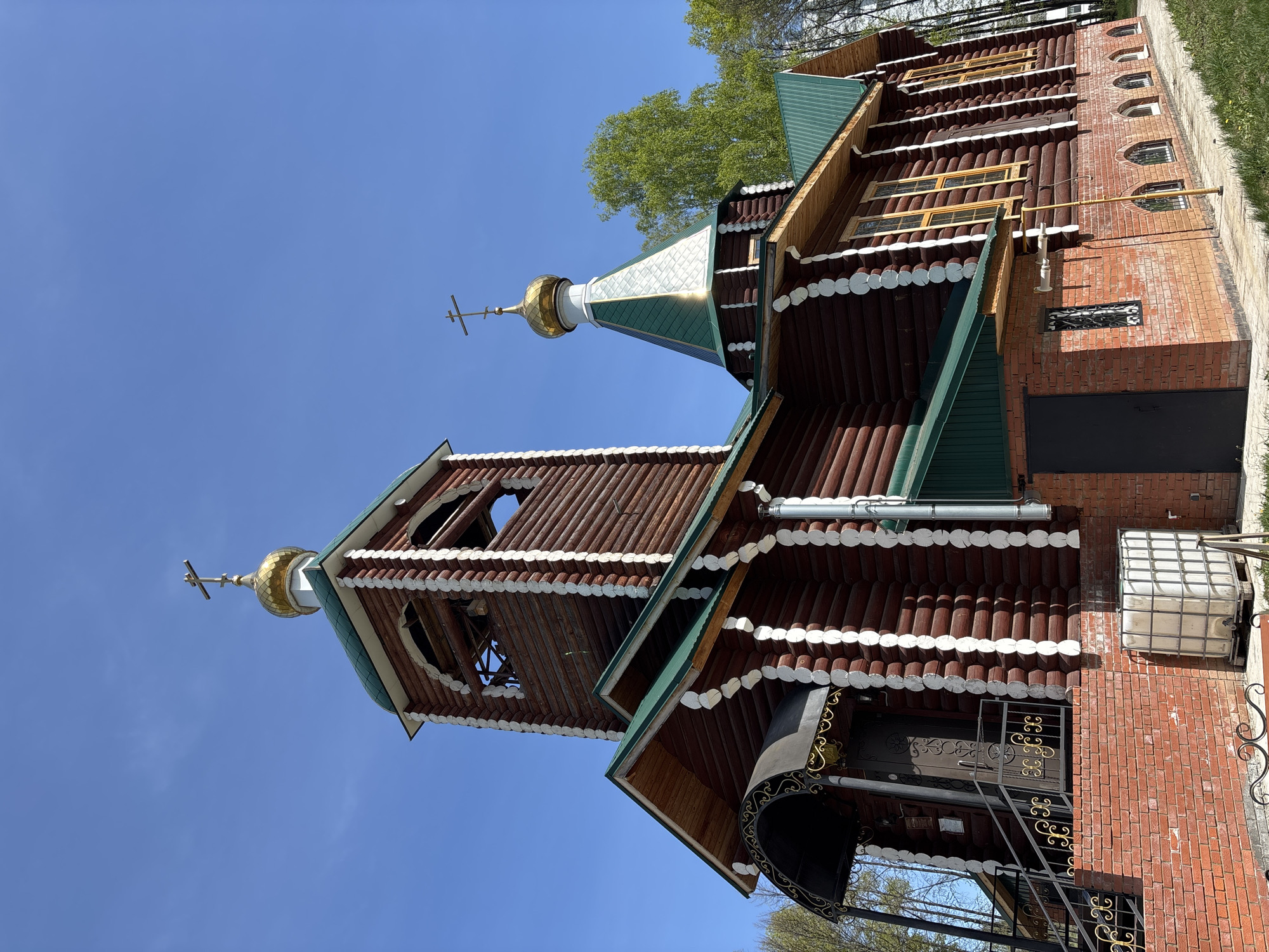 The image shows a wooden Russian Orthodox church in the city of Chaikovsky, Russia. The church has a main section built from horizontal logs and a brick section. The main section has dark brown logs with white decorative elements and a green roof. The church features two towers topped with golden domes and Orthodox crosses. The sky is clear and blue. A metal staircase leads to a decorated doorway. An outdoor air conditioning unit or water tank is attached to the brick portion of the building. There are trees visible in the background.