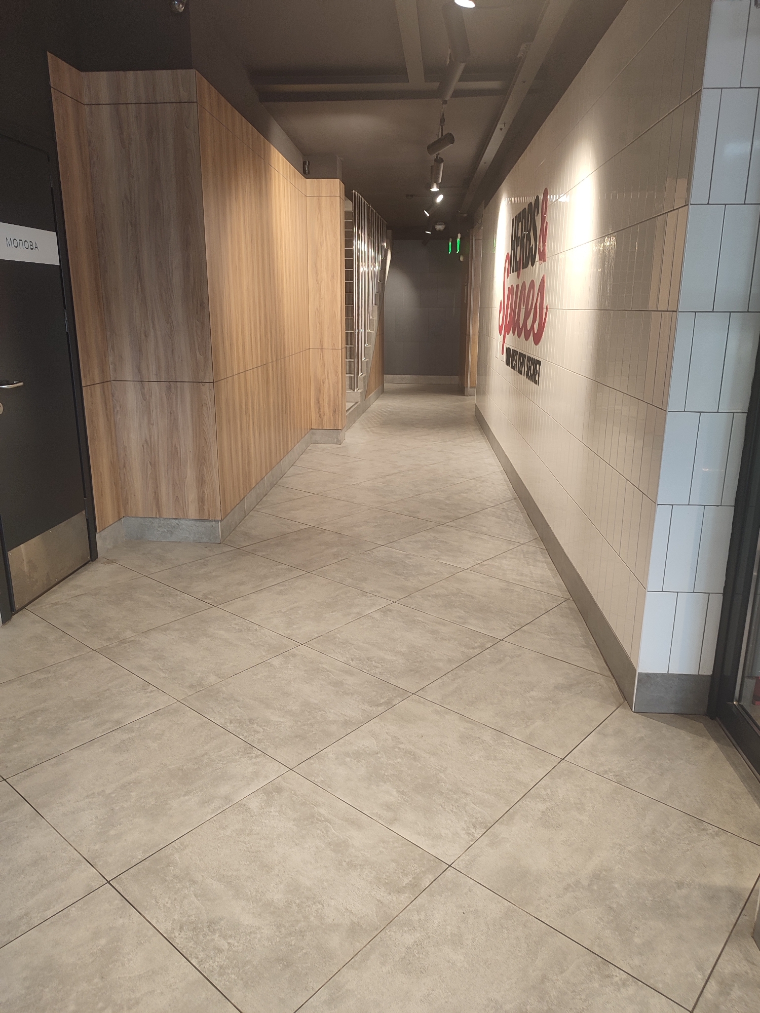 The image depicts an interior view of a hallway or corridor. The left side of the hallway features a dark door with a metallic handle and a white label with Cyrillic text, likely "МОЛОВА," which translates to "Molova" or "Milk." Adjacent to the door is a wall clad in vertical wooden panels. The right side of the hallway is lined with a tiled wall, predominantly white, with red lettering spelling out "HERBS & SPICES" and a smaller tagline below. The floor is tiled with large, square, light-colored tiles laid in a diamond pattern. The ceiling is dark and appears to have recessed lighting fixtures. In the distance, the hallway continues, with a railing and stairs visible on the right. The overall impression is of a modern, clean, and well-lit commercial or public space. There are no people visible in the image. This scene is not in Chernivtsi, Ukraine.