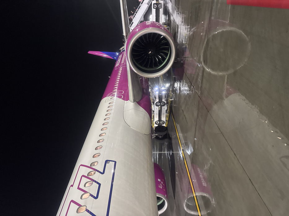 A low-angle, rotated view captures a Wizz Air Airbus on a wet tarmac at night. The fuselage, with its distinctive magenta and white livery, runs vertically alongside a powerful jet engine. Reflections distort the image, creating circular patterns on the pavement. Landing gear with bright lights and passenger stairs add to the dynamic composition. The tail fin and company logo are visible, emphasizing the plane's identity against the dark background.