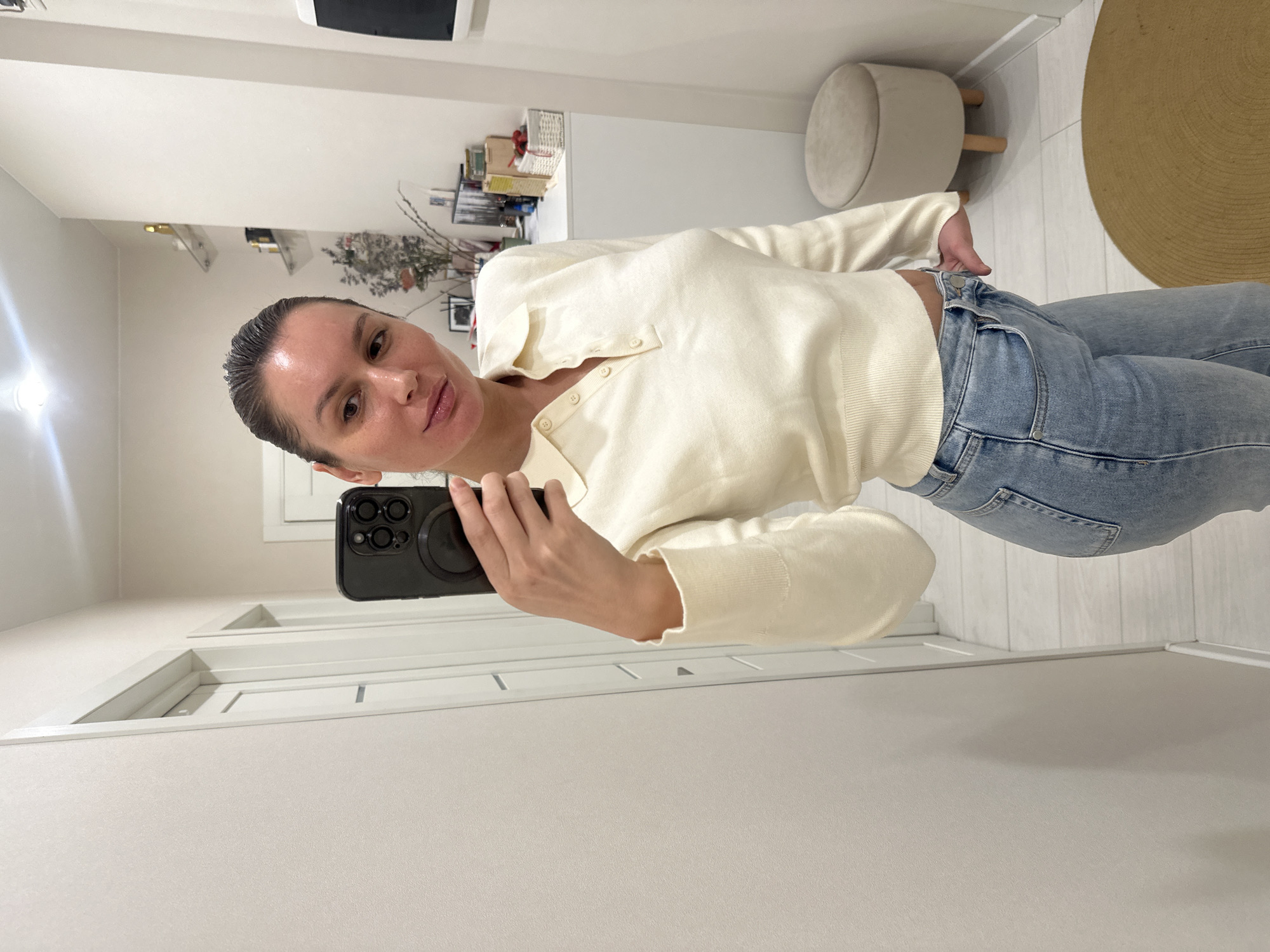 The image captures a woman taking a mirror selfie indoors. She stands slightly off-center, facing the viewer with a neutral to slightly pensive expression and subtly pouting lips. Her hair is pulled back and appears wet or slicked, and her skin has a slight sheen.
She is wearing a light cream or off-white long-sleeved knitted top with a button-up placket and light blue denim jeans. Her right hand holds a black smartphone with a prominent camera module and a round MagSafe-like attachment, pointed towards the mirror to capture the shot. Her left hand is casually tucked into her jeans.
The background depicts a clean, modern interior space, likely a room or hallway within a residence. The walls are white, and the floor is light-colored, possibly wood or laminate. Visible furniture and decor include a small, round, light-colored upholstered stool with wooden legs, and a large, round natural fiber mat on the floor. In the background, there are shelves or surfaces holding various items like baskets, books, and dried floral arrangements. A white door frame is also visible. The lighting is artificial, suggesting an indoor setting, potentially in the evening. There is no visible text in the image. The overall impression is one of a casual, everyday moment in a tastefully decorated home in Moscow, Russia.