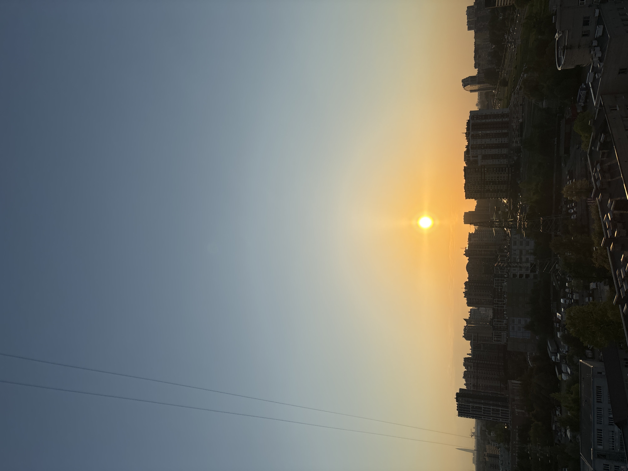 The image depicts a cityscape at sunset. The sun is low on the horizon, casting a warm orange glow across the sky and silhouetting the buildings. The city appears to be densely populated with a mix of high-rise buildings and smaller structures. The perspective is from an elevated position, possibly a high-rise building or a hill. There are power lines visible in the lower-left corner of the image. The city and exact location are Unknown.