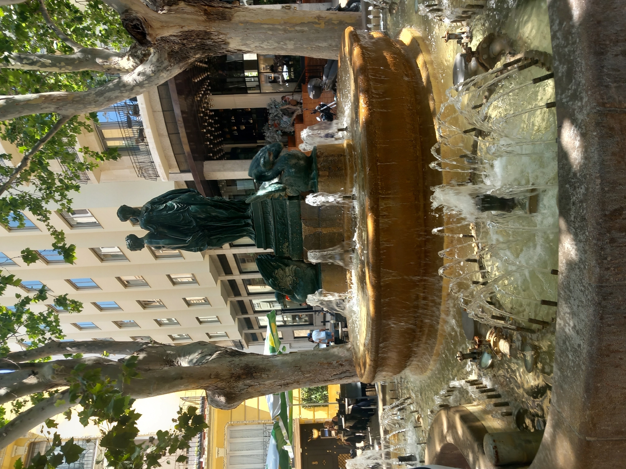 The image depicts a fountain in the city of València, Spain, located in a public square lined with buildings and trees. The fountain is circular with multiple tiers, featuring bronze sculptures of human figures and winged creatures. Water sprays from various spouts, creating a dynamic and refreshing scene.
In the background, a multi-story building with a modern architectural style dominates the skyline. The building has numerous windows and balconies, suggesting it may be a hotel or residential complex. Adjacent to it, a cafe or restaurant with outdoor seating and umbrellas is visible, with a few patrons seated at tables. The presence of a motorcycle parked near the fountain suggests an urban setting.
The trees in the foreground are mature with broad trunks and lush green foliage, providing shade to the area. The lighting suggests it is a sunny day, with shadows cast by the trees and buildings. The overall atmosphere is lively and inviting, indicative of a bustling urban square.