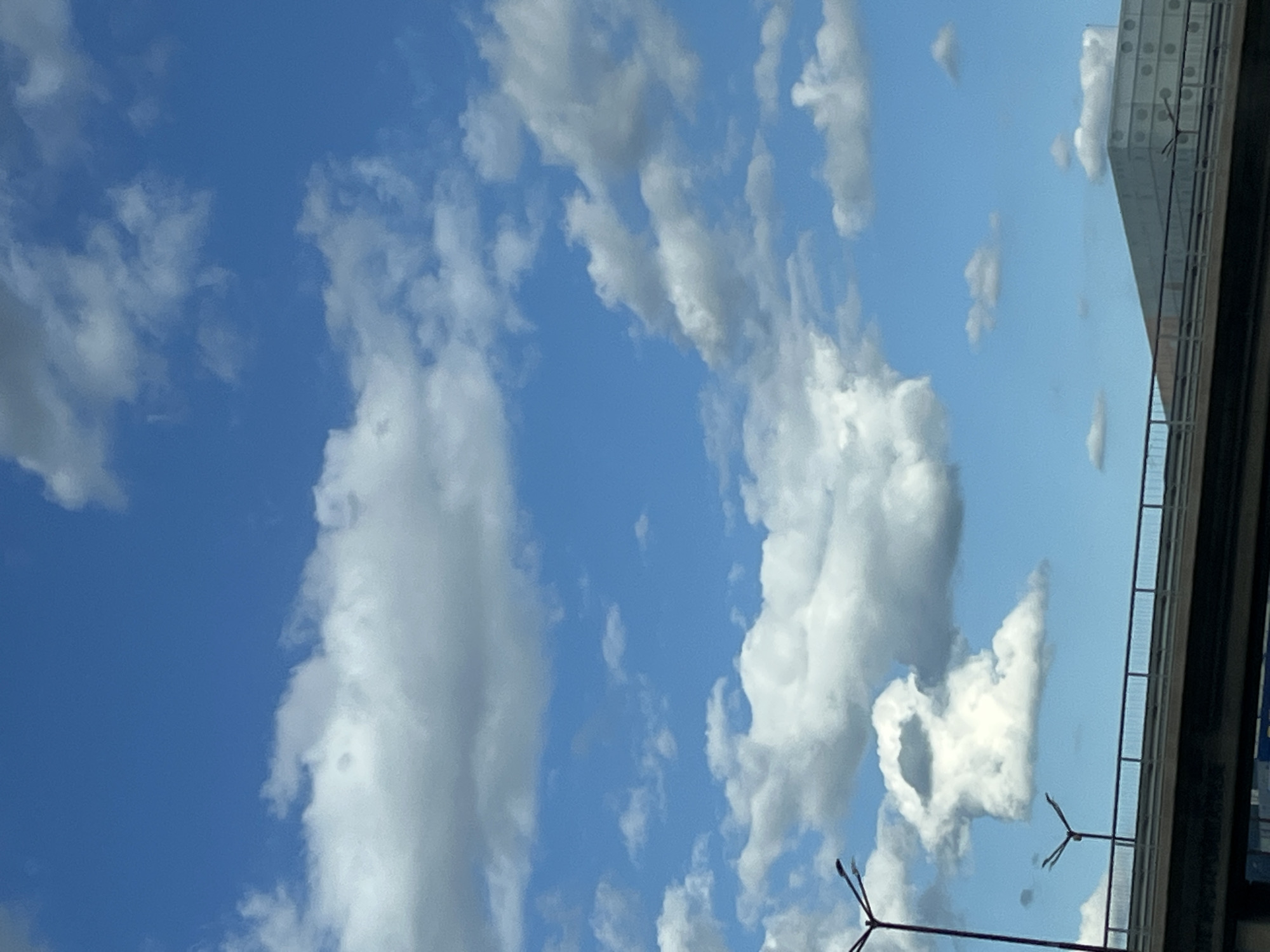An outdoor, eye-level, vertical, medium shot captures a bright, sunny day with scattered white clouds against a vibrant blue sky. In the foreground, the top of a dark, elevated roadway structure is visible, with a metal railing along its edge. Two streetlights with multiple arms are positioned on the left side of the roadway. To the right, a modern, multi-story building with a distinctive facade featuring circular patterns is partially visible behind the roadway. The scene appears to be in an urban environment, possibly a city, given the presence of the roadway and the building. The lighting suggests daytime.