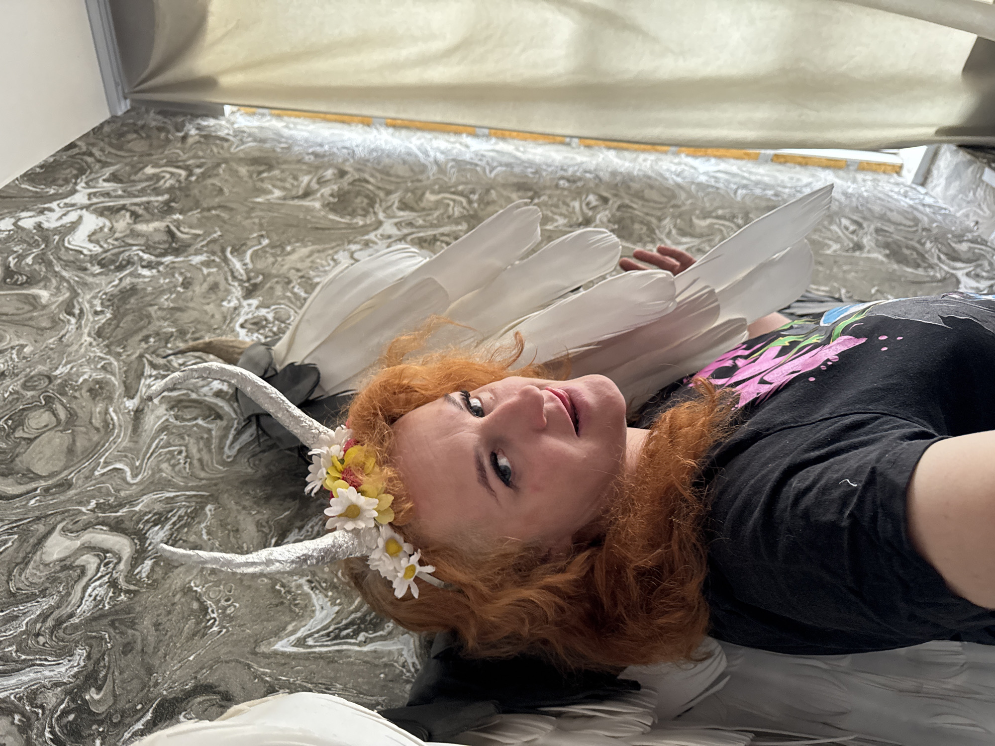 A red-haired person wearing a black t-shirt with pink designs, a floral headpiece, and white horns lies on a marbled surface under a light-colored awning. White feathers surround the person's head and body. The location in the city of Chaikovsky, Russia cannot be verified from the image.