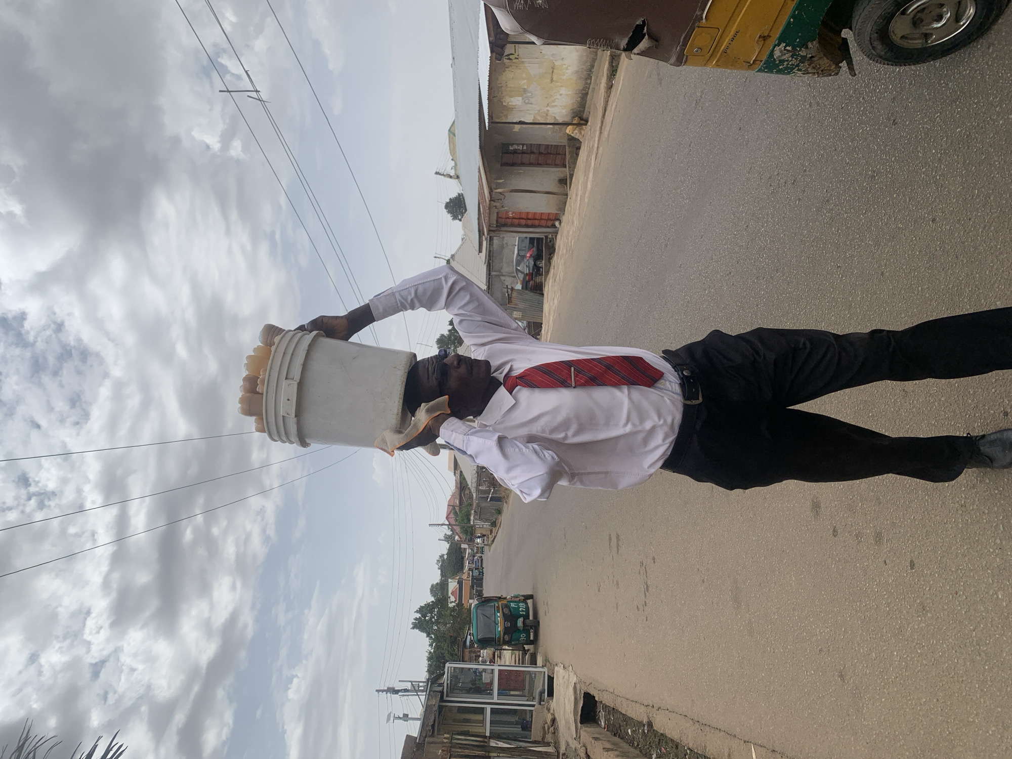 A man in a white shirt and red tie balances a large bucket on his head while walking on a paved road in Jos, Nigeria. He wears black pants and shoes. The bucket appears to be filled with food. In the background, there are buildings and a three-wheeled vehicle. Overhead, there are electrical wires against a cloudy sky. The road is mostly empty.