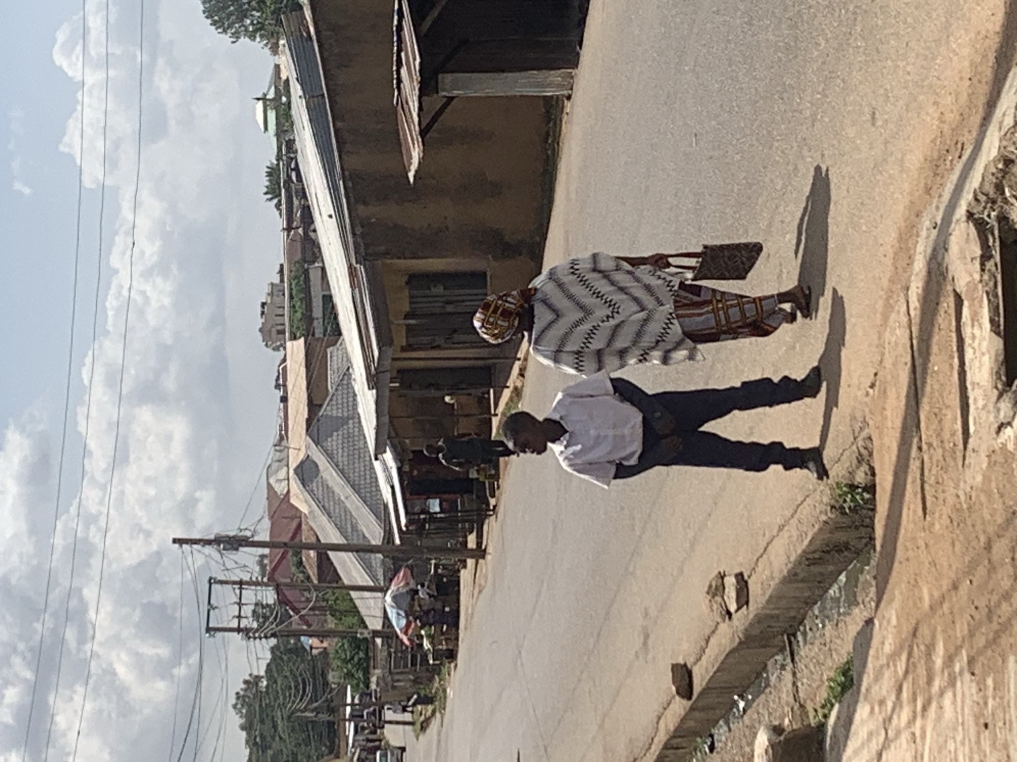 This media file captures an outdoor street scene, likely in a town or city in Jos, Nigeria, given the provided context. The image shows two main adult figures walking along an asphalt road under a bright, cloudy sky, suggesting daytime, possibly mid-morning or afternoon due to the visible shadows.
On the right side of the street, a woman walks away from the camera. She is dressed in a patterned garment, possibly a dress or long top, featuring a white and black zig-zag or geometric design, and wears a matching or similarly patterned head covering. She carries a dark handbag over her right shoulder. To her left and slightly closer to the camera, a man is also walking, seen in profile. He is wearing a white long-sleeved shirt and dark trousers.
The street is lined with buildings of varying architecture, some appearing to be shops or residences, with corrugated metal elements visible on their roofs or awnings. In the background, further down the street, other indistinct figures can be seen. On the left side of the road, utility poles with numerous power lines crisscross the frame, indicating an urban infrastructure. Along the right edge of the street, a partially visible open drainage channel or gutter runs parallel to the road. No legible text is visible in the image. The overall scene depicts a mundane moment of daily life and transit in a bustling street environment.