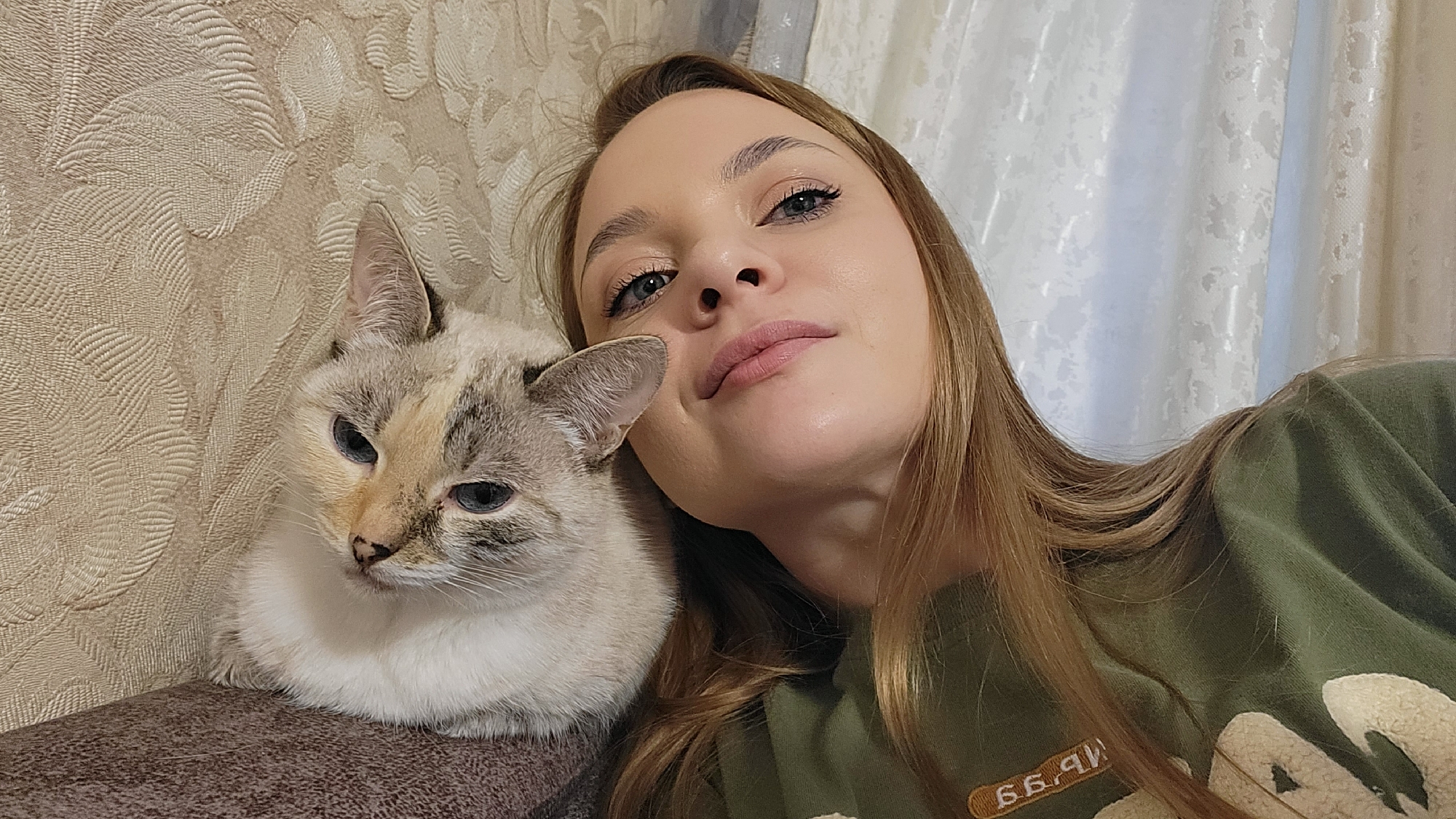 The image shows a woman with long brown hair taking a selfie with a light-colored cat with blue eyes. The cat is resting on a brown surface, likely part of a sofa. The woman is wearing a green sweater. The background includes a patterned wall and a white curtain. The location is Chaikovsky, Russia, based on metadata.