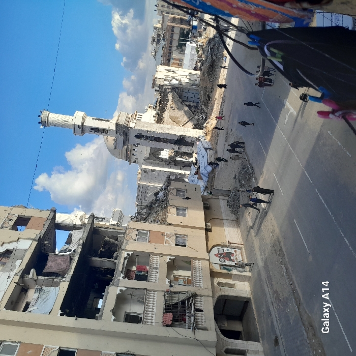 This image displays a street scene in Khan Yunis, Palestinian Territory, marked by extensive structural damage. On the left, a multi-story building exhibits heavy destruction, with multiple floors exposing interiors, missing walls, and hanging debris. Laundry is visible on balconies and exposed sections of the building. A paved two-lane road with white lane markings occupies the midground. Approximately 20 to 30 individuals are seen walking and standing in small groups on the street and amidst rubble. In the background, a large mosque is severely damaged, its minaret leaning but largely intact amidst collapsed sections of the main structure and surrounding debris. Additional damaged buildings and some white tent-like structures are discernible further in the background. The sky is blue with scattered white clouds. A clothesline with dark fabric and colorful clothespins is partially visible in the extreme right foreground.